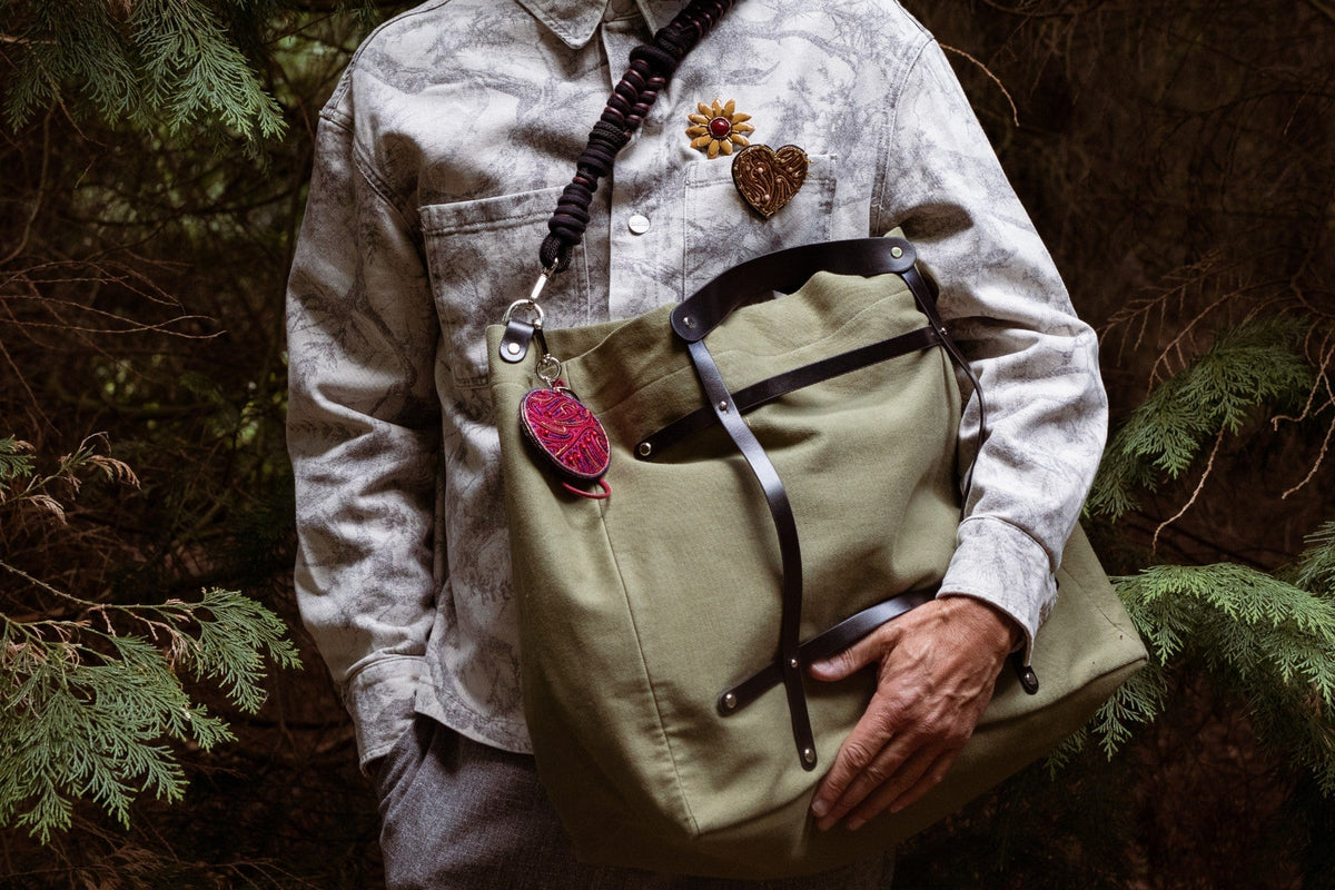 Person wearing a light-colored shirt and green bag with an embroidered burgundy round NZ with nature-themed decorations, standing in a forest setting.