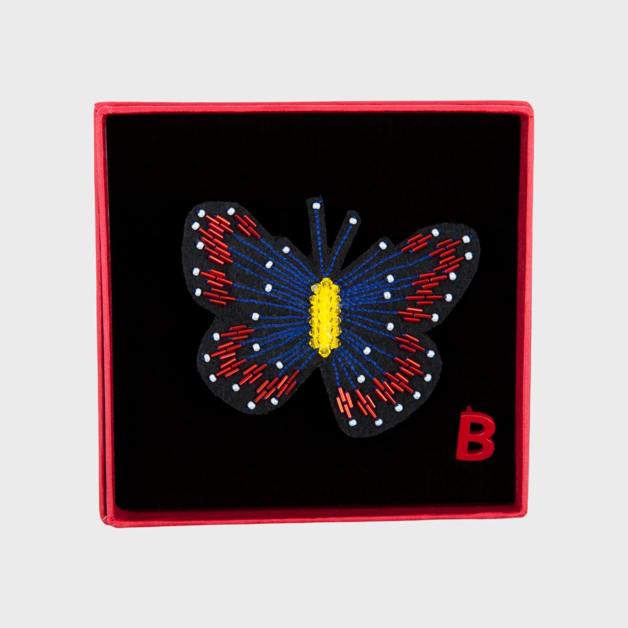 An embroidered brooch representing a black butterfly with blue and red details in a box. 