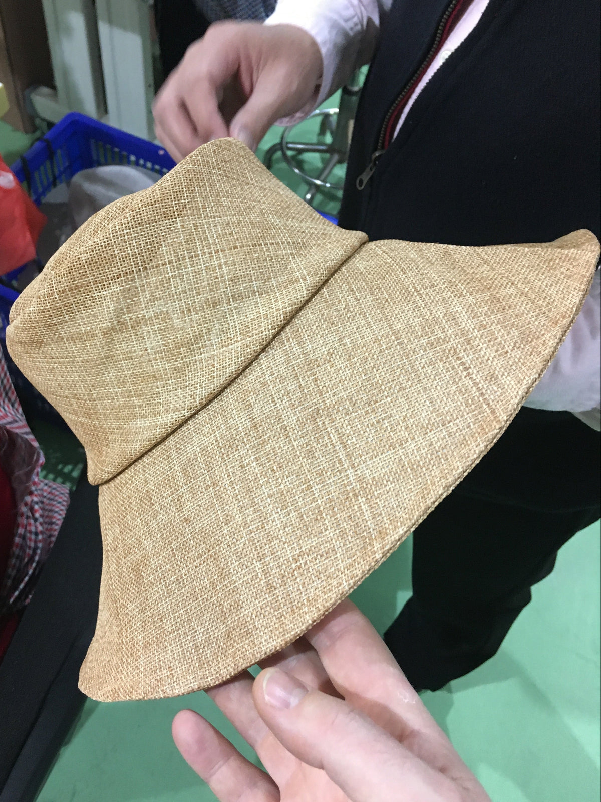 A light tan woven bucket hat being held by hands in what appears to be a workshop or store setting, with a person in a black vest visible in the background.