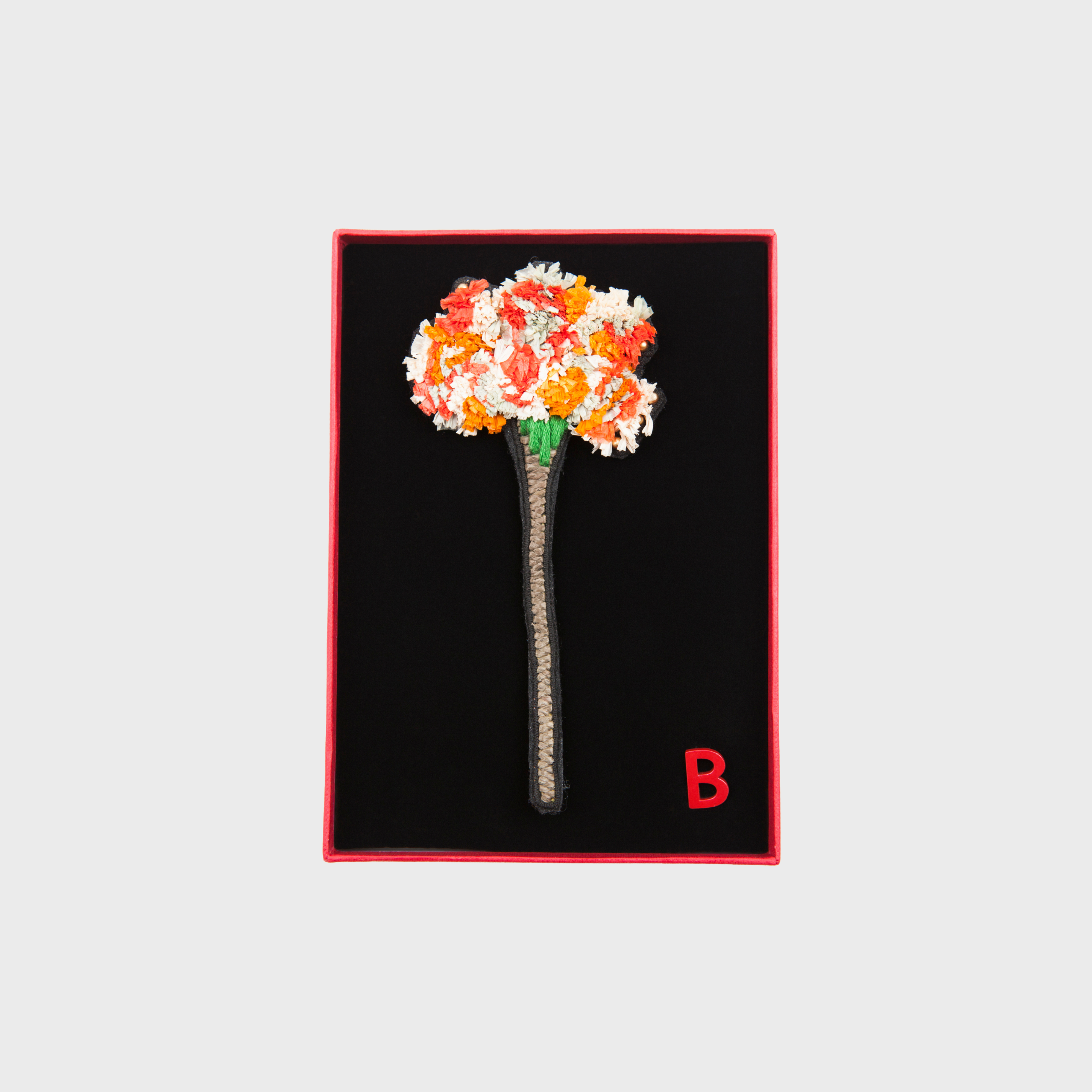 An embroidered brooch representing a long flower with orange and white details in a box. 