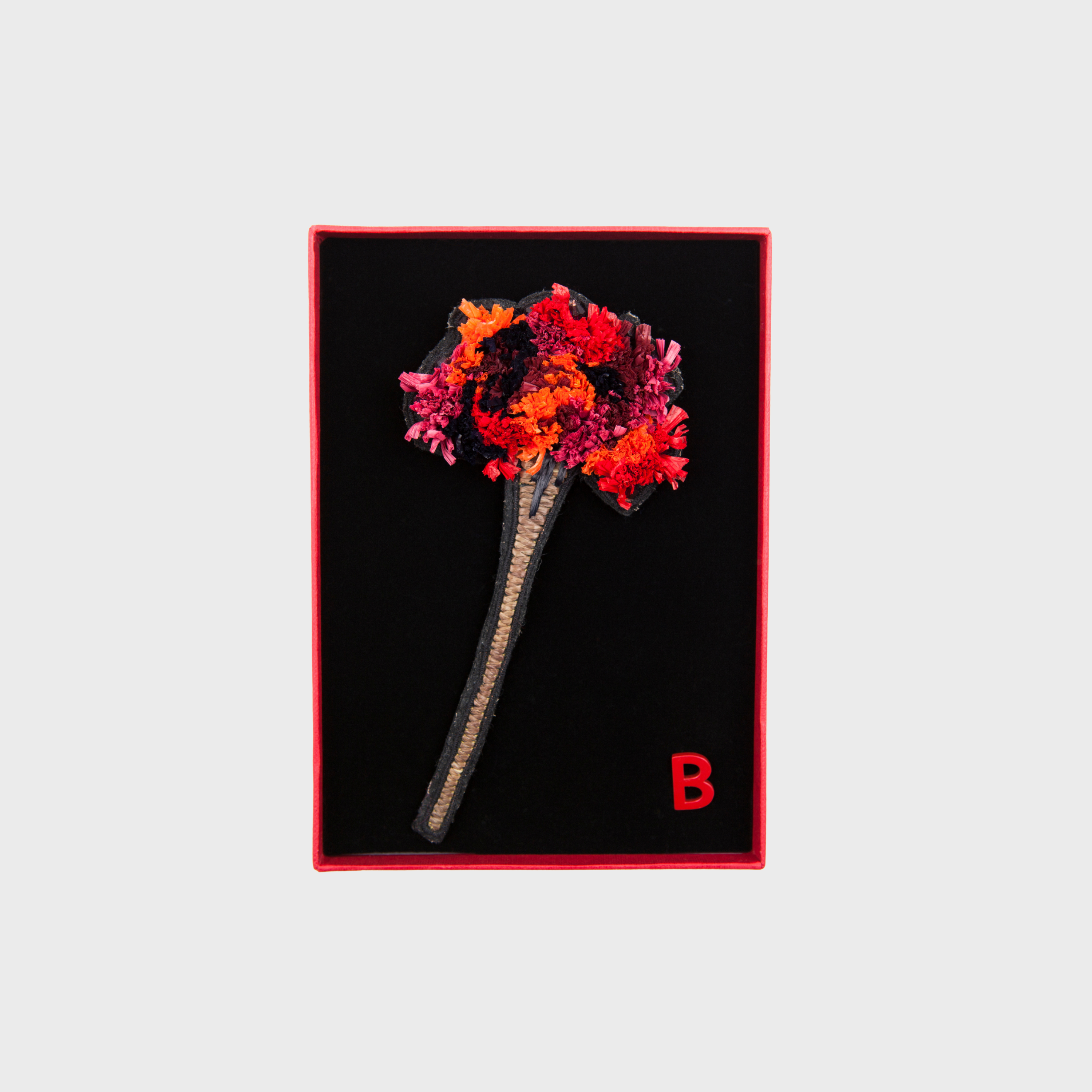 An embroidered brooch representing a long flower with red , orange and brown details in a box. 