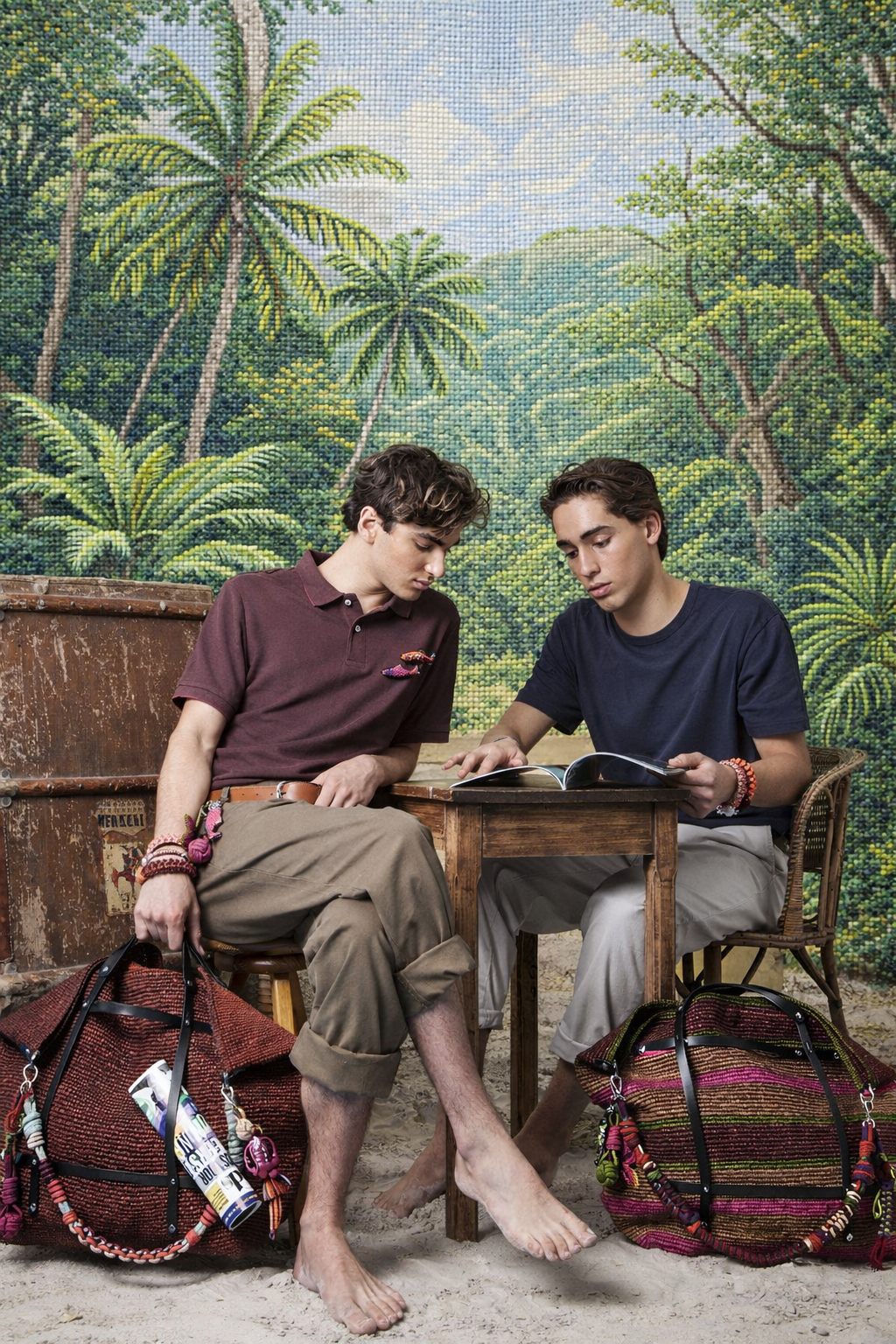 Two men sitting and reading at a table with a tropical mural in the background both carrying brown and striped brown, red, and green candides weekender bags.