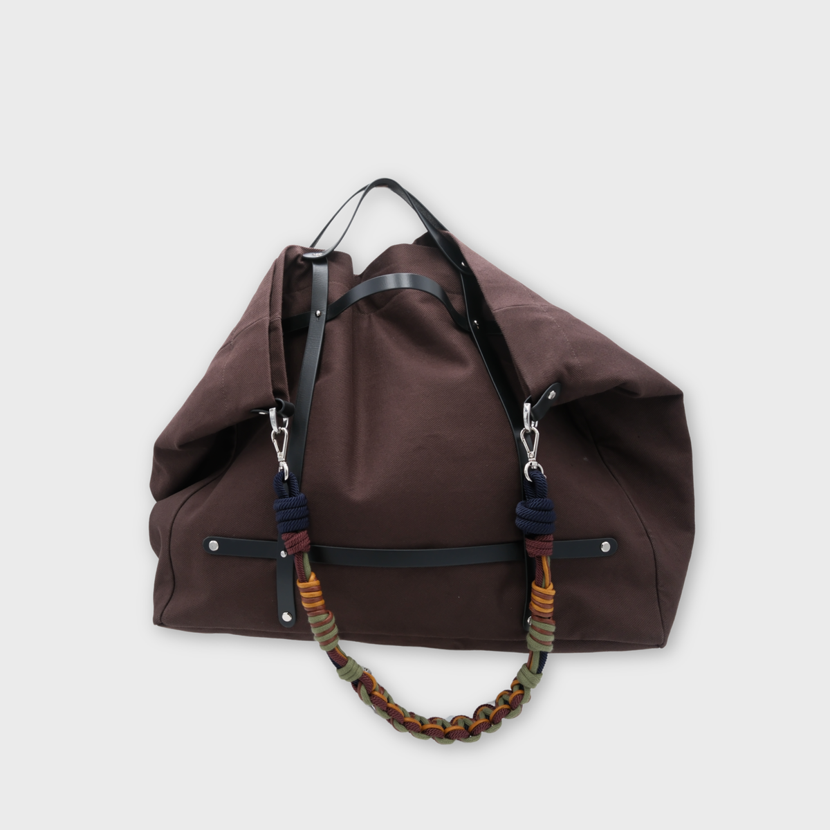 Model carrying a chcolate canvas weekend or duffle bag with a black leather harness, paired with a neutral toned shoulder strap in yellow, green and brown.