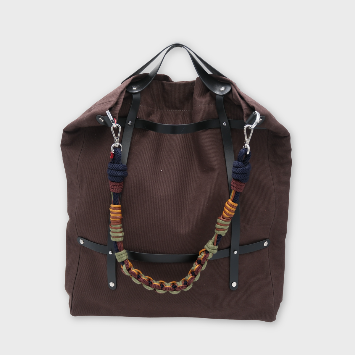 A canvas tote bag in a chocolate color, featuring brown leather straps and a decorative shoulder strap with blue, green, yellow details. 