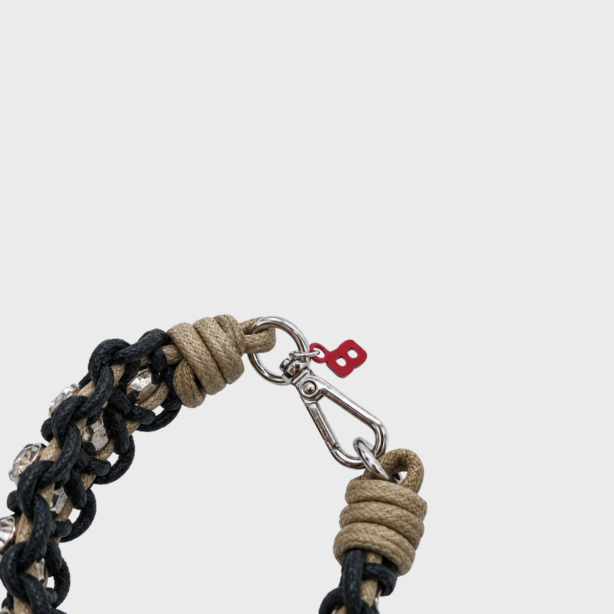Close-up of a woven bracelet with black and beige braided cords, a silver metal clasp, and a small red tag on a light gray background.