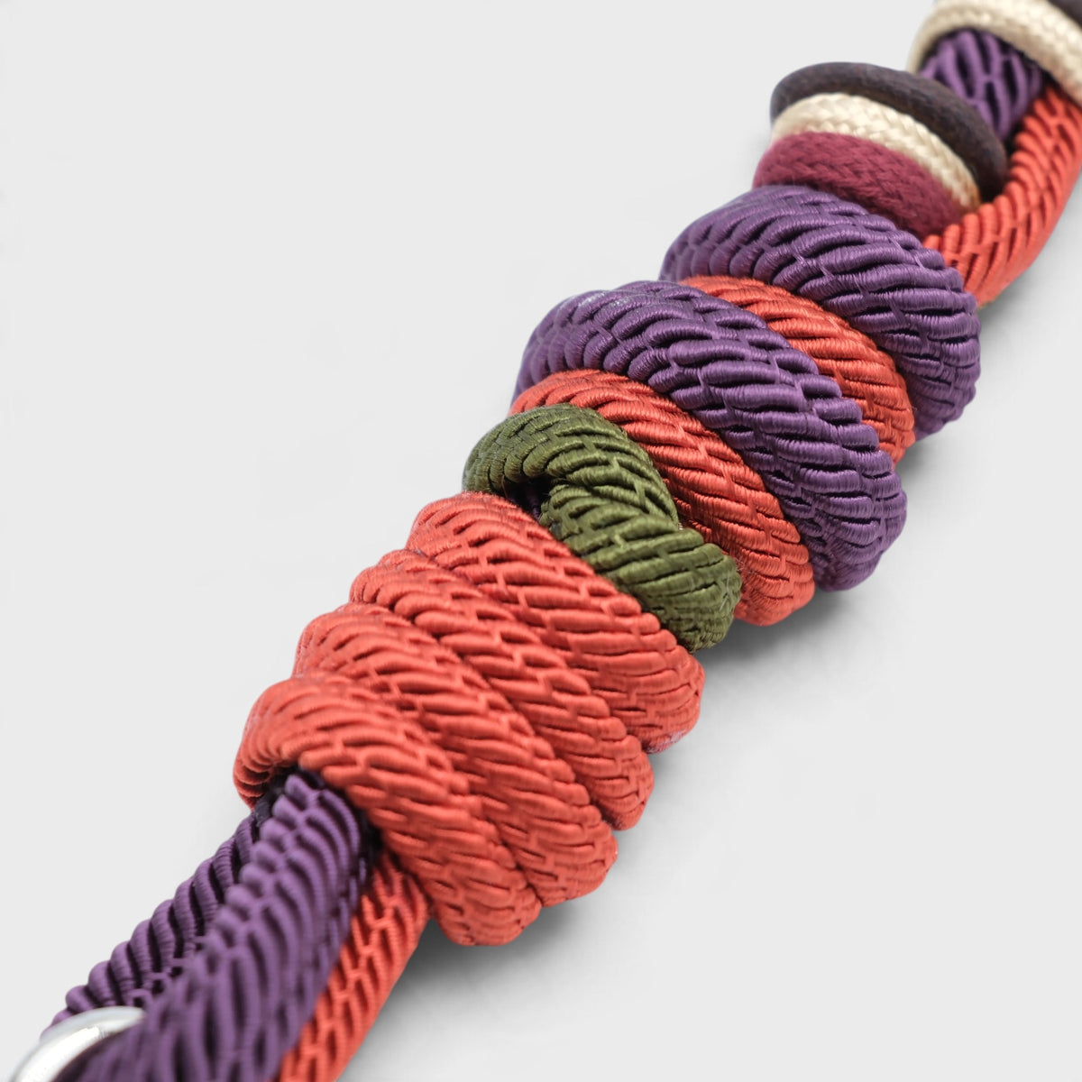 A close-up of a braided macramé bag strap with knotted detailing, shown diagonally on a light gray background. The cord features multiple colors including purple, coral red, olive green, and cream.