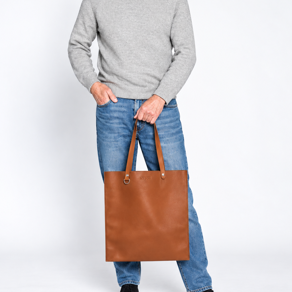 A smiling older man wearing a gray turtleneck sweater, blue jeans, and black shoes stands against a white studio background, holding a large brown leather tote bag.