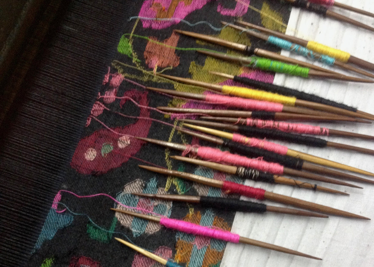 A close-up of a handwoven textile with a colorful floral pattern on a dark base, shown alongside multiple wooden knitting needles wrapped with bright pink, green, yellow, and blue thread.