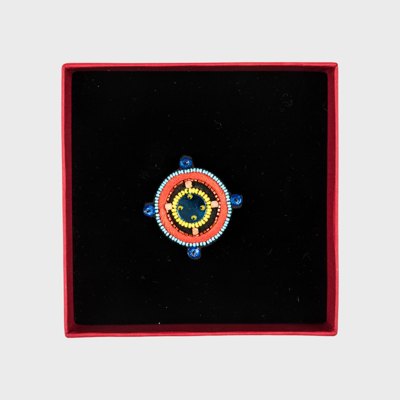 An embroidered brooch representing a compass with blue and red details in a box. 