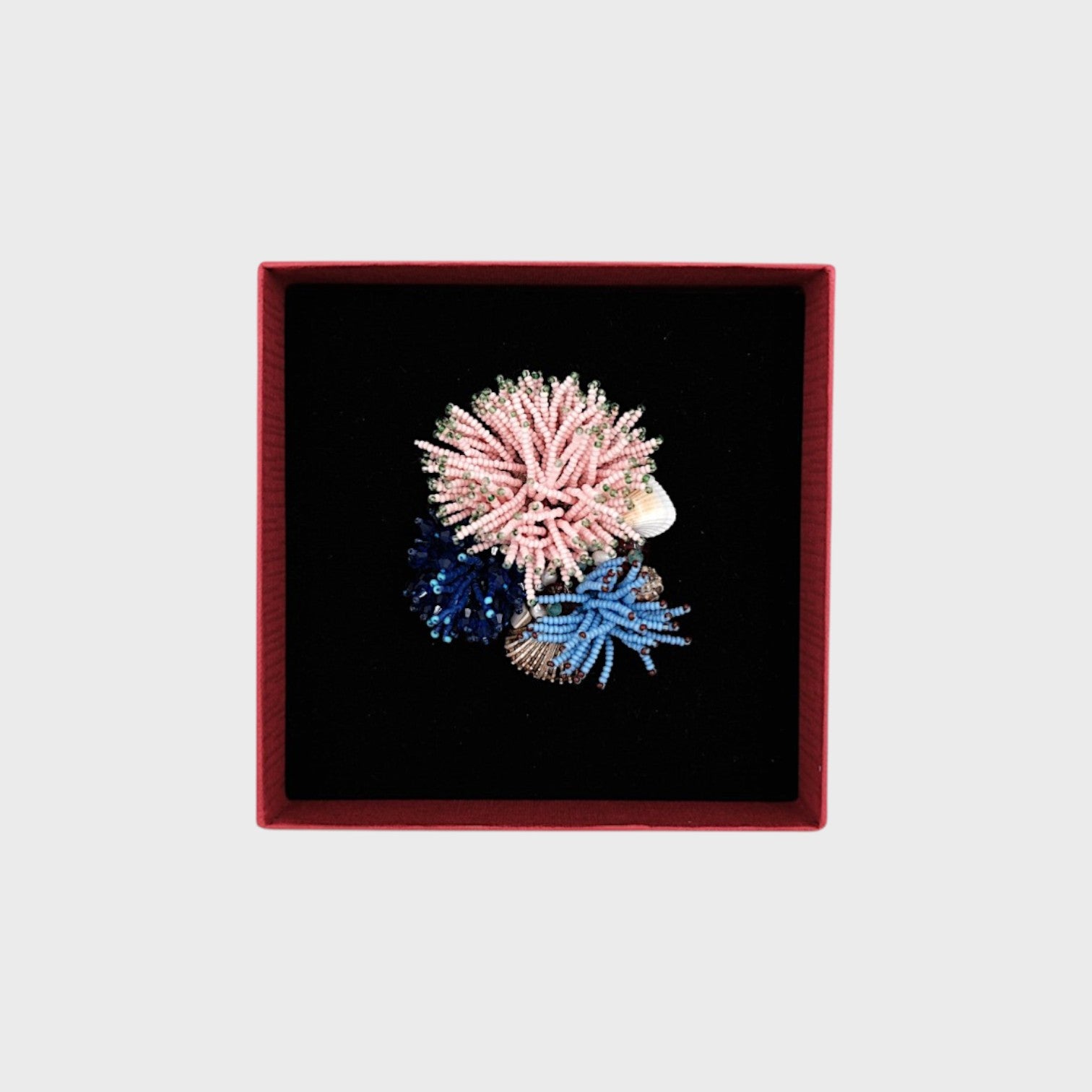 A embroidered coral brooch with pink, dar blue and blue details in a box