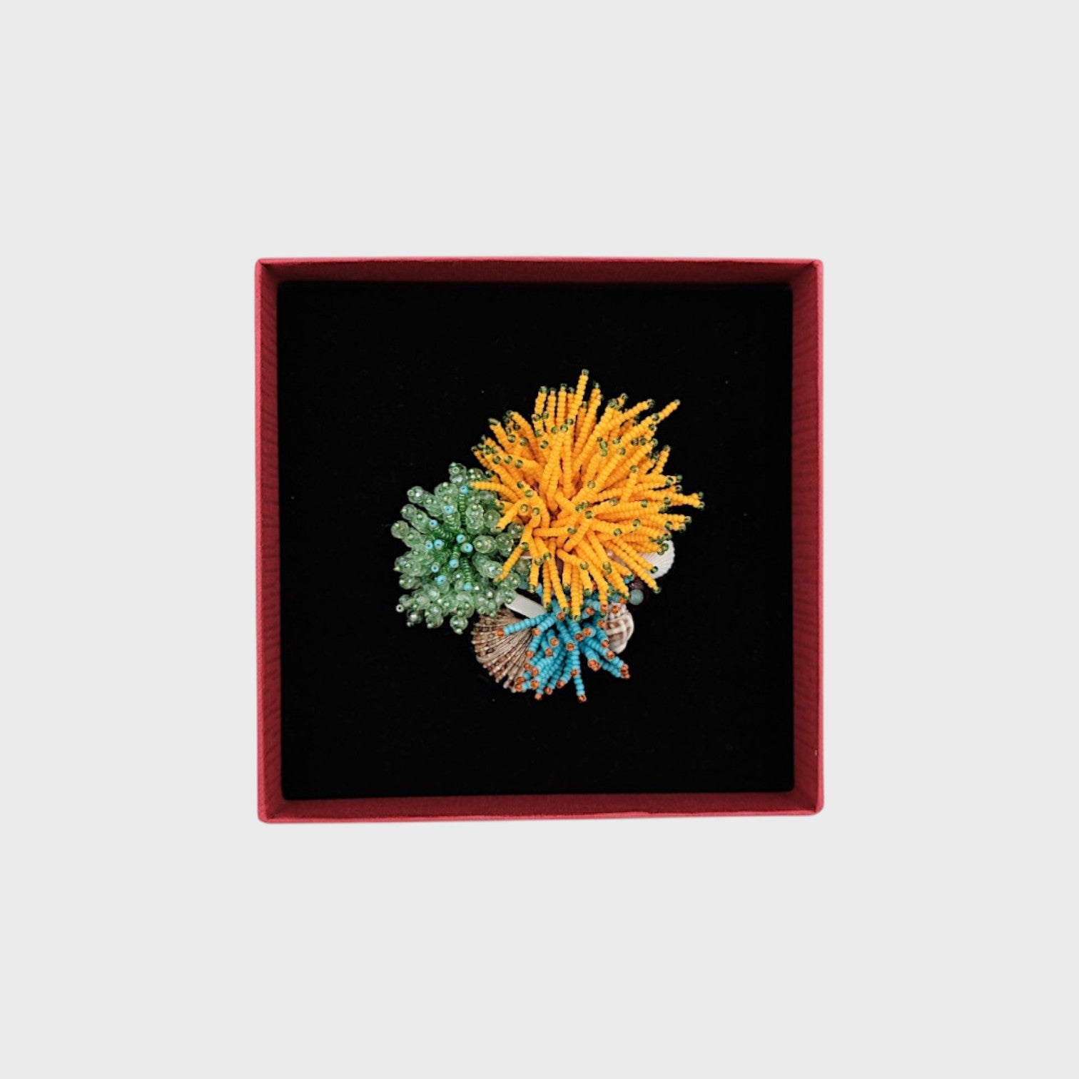 A coral embroided brooch with yellow, green and blue details in a box. 