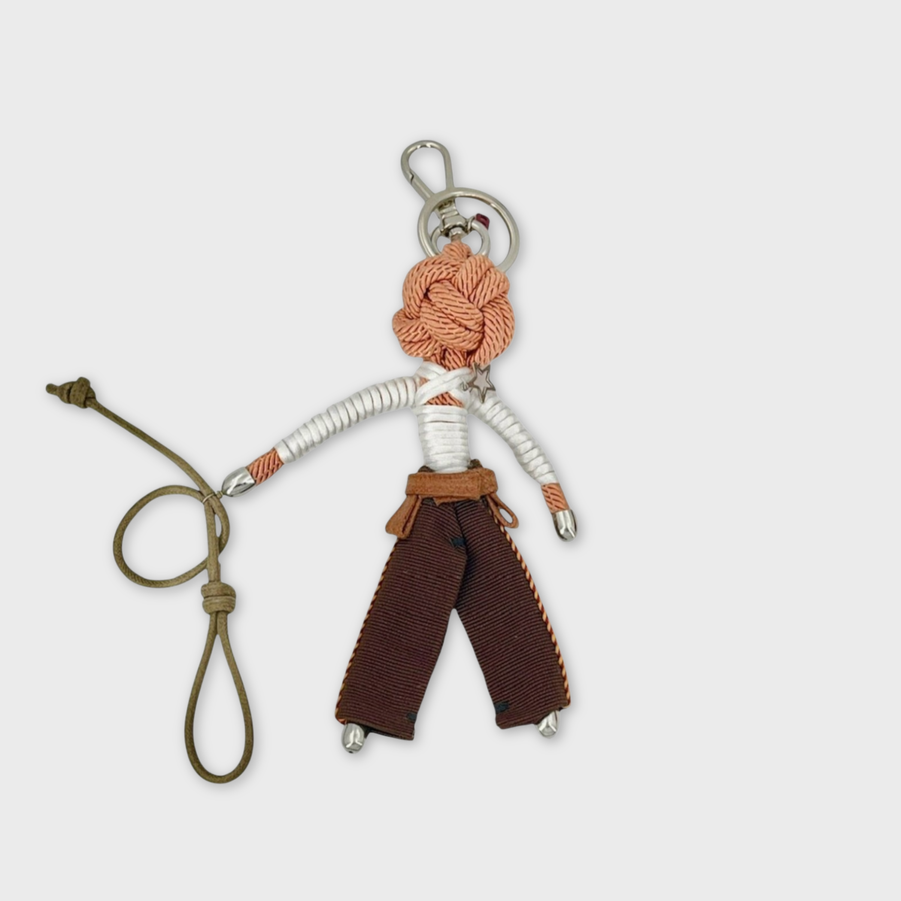 Handmade tan macramé bag charm featuring a male character, meant to be a cowboy with his lasso, wearing traditional brown cowboy trousers, a belt and a long sleeves white top. He is attached to a silver clasp.