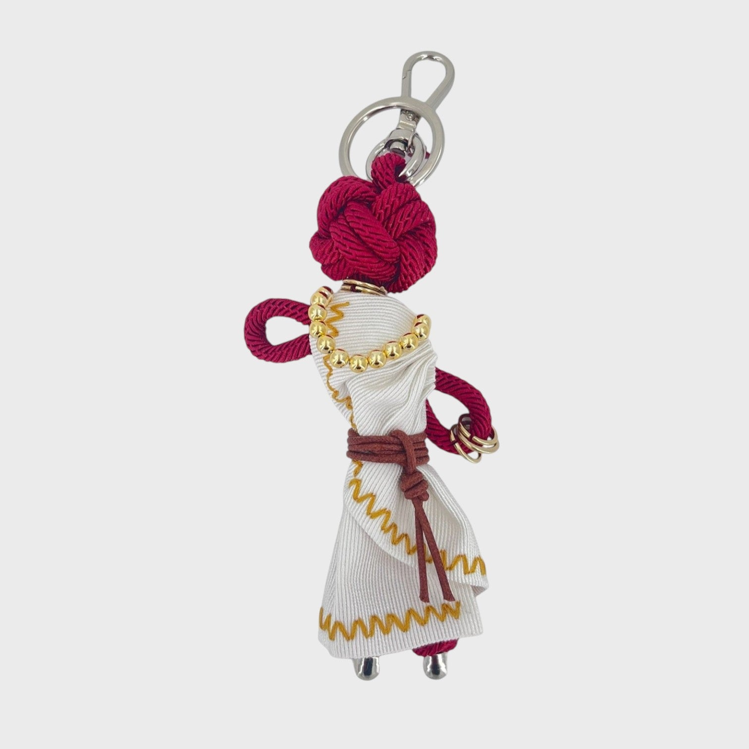 Handmade burgundy macramé bag charm meant to represent a woman deity wearing a maxi white wrap dress with yellow trimming, a brown belt wrapped around her waist, golden beaded necklace and a double golden bracelet around her wrist. She is attached to a silver clasp.