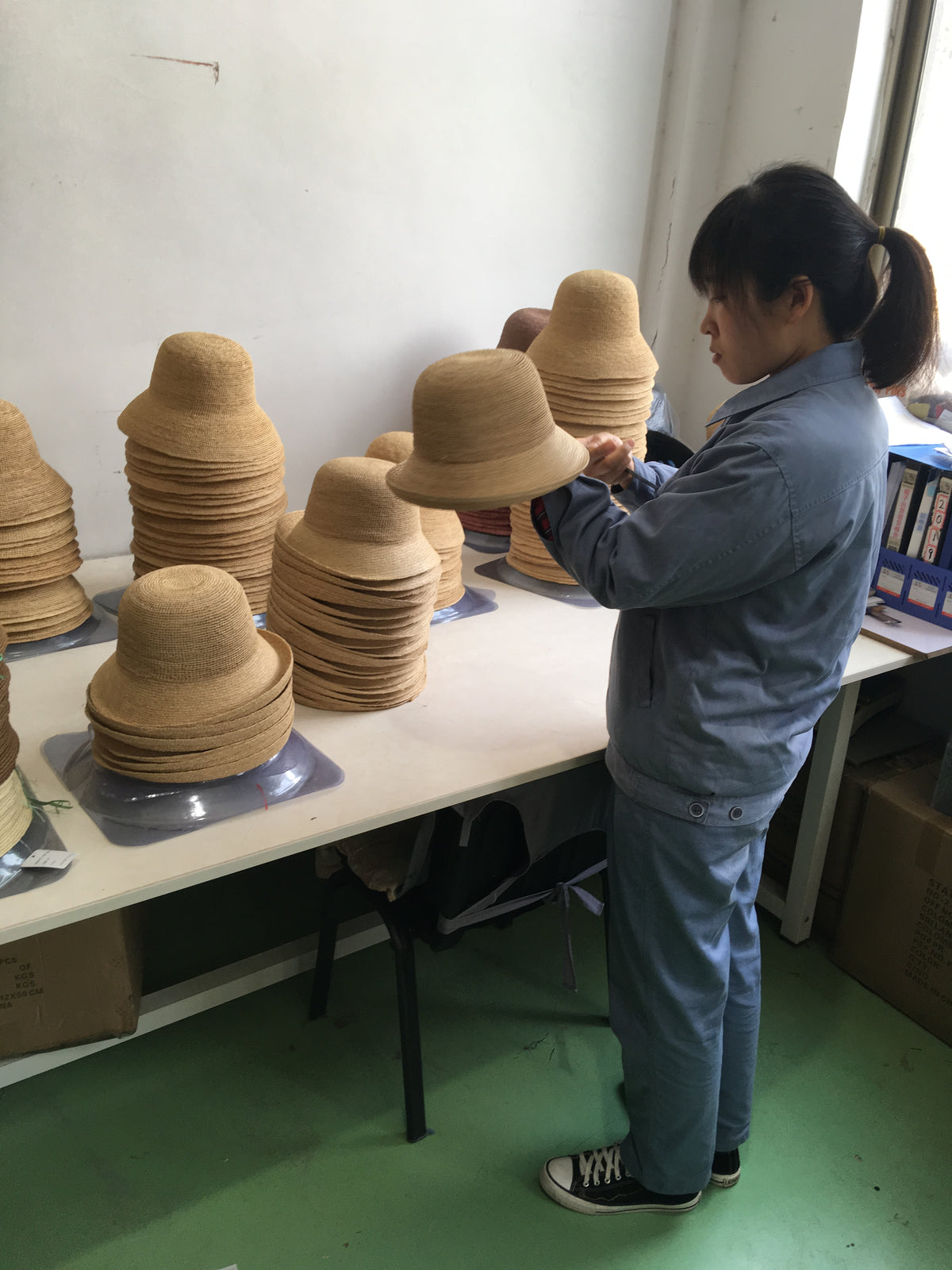 A woman in a blue uniform stands at a table handling a straw bob, with stacks of similar straw hats arranged on the table in a workshop setting.