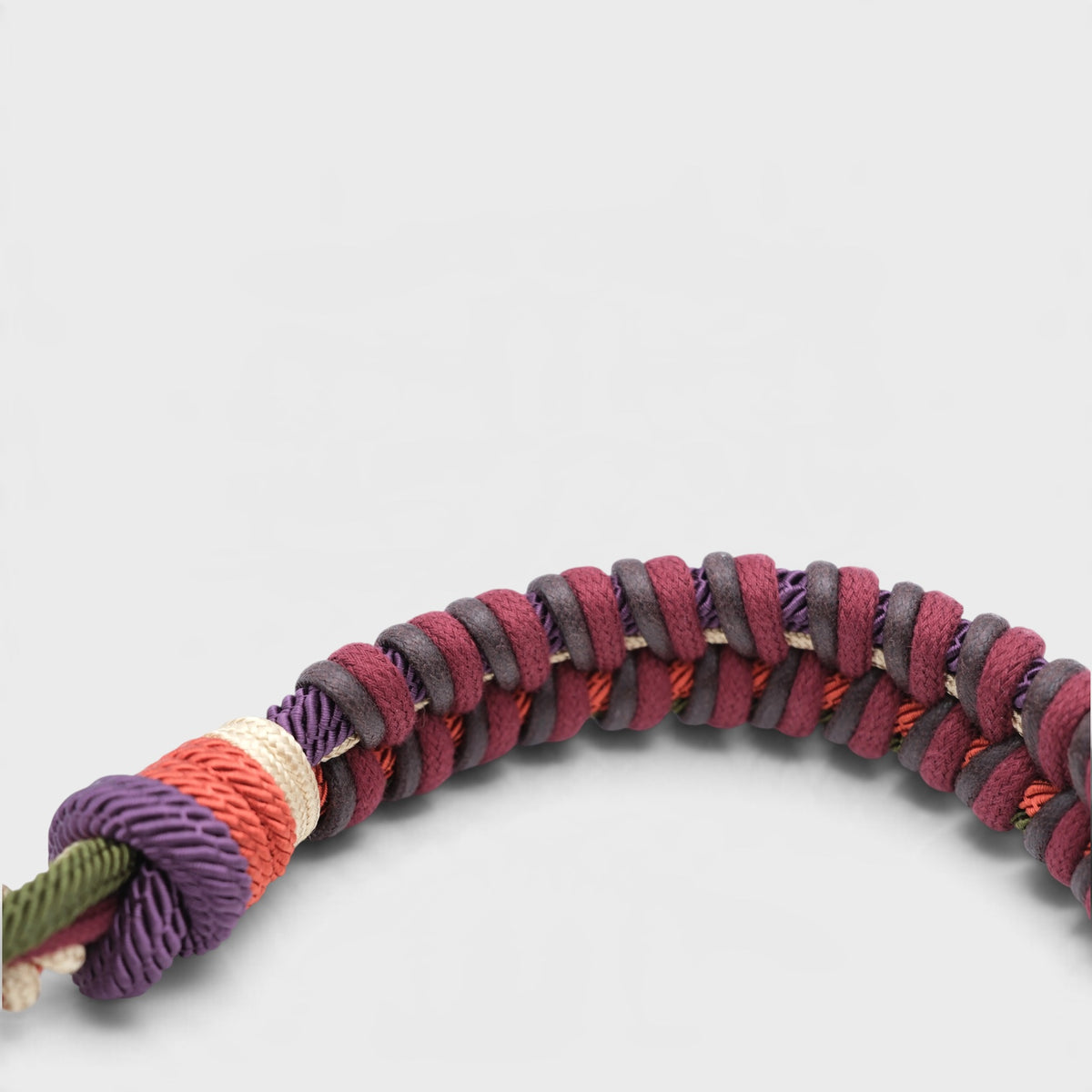 A close-up of a braided macramé bag strap, featuring alternating bands of burgundy, dark brown, and purple, with a knotted accent in purple, cream, and orange, and a green cord end visible on the left, set against a light gray background.