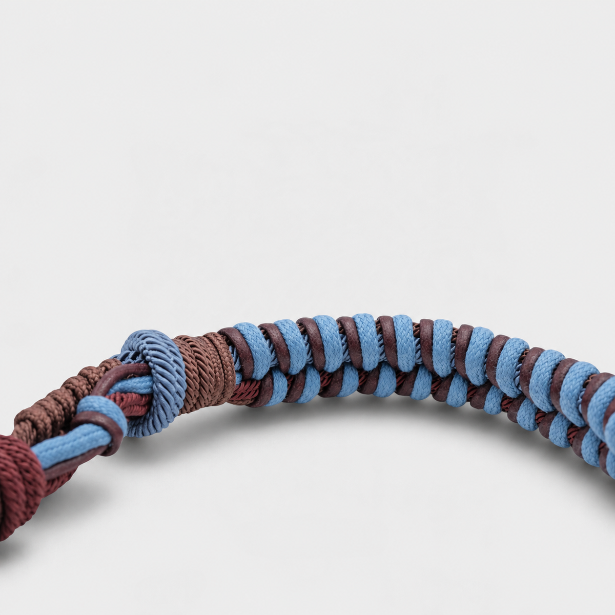 A close-up of a braided macrame bag strap, featuring alternating light blue and dark brown braided loops with a knotted end, set against a plain light gray background.