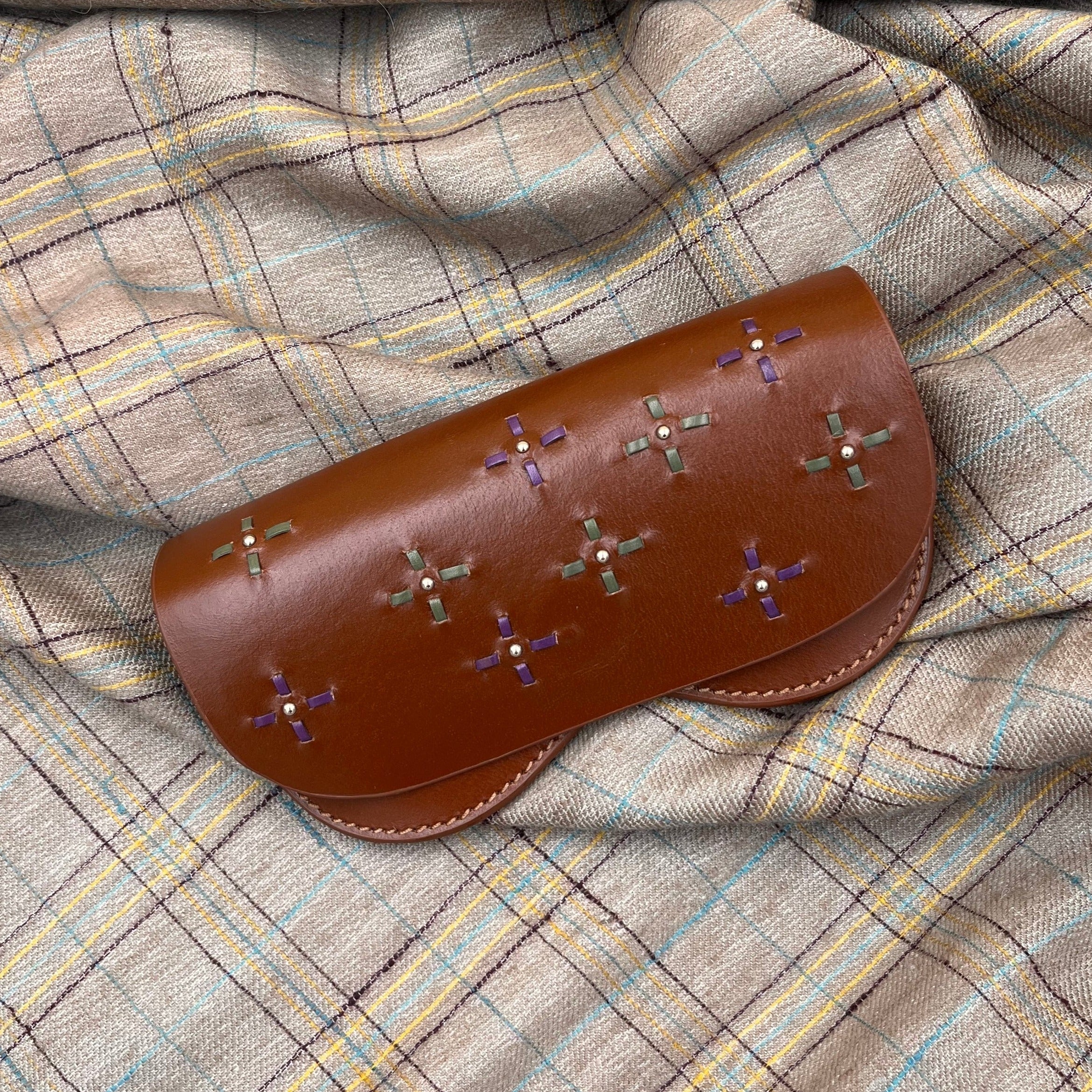 A brown leather dots glasses case with green and purple symbols embroidered on it on a grey blanket. 