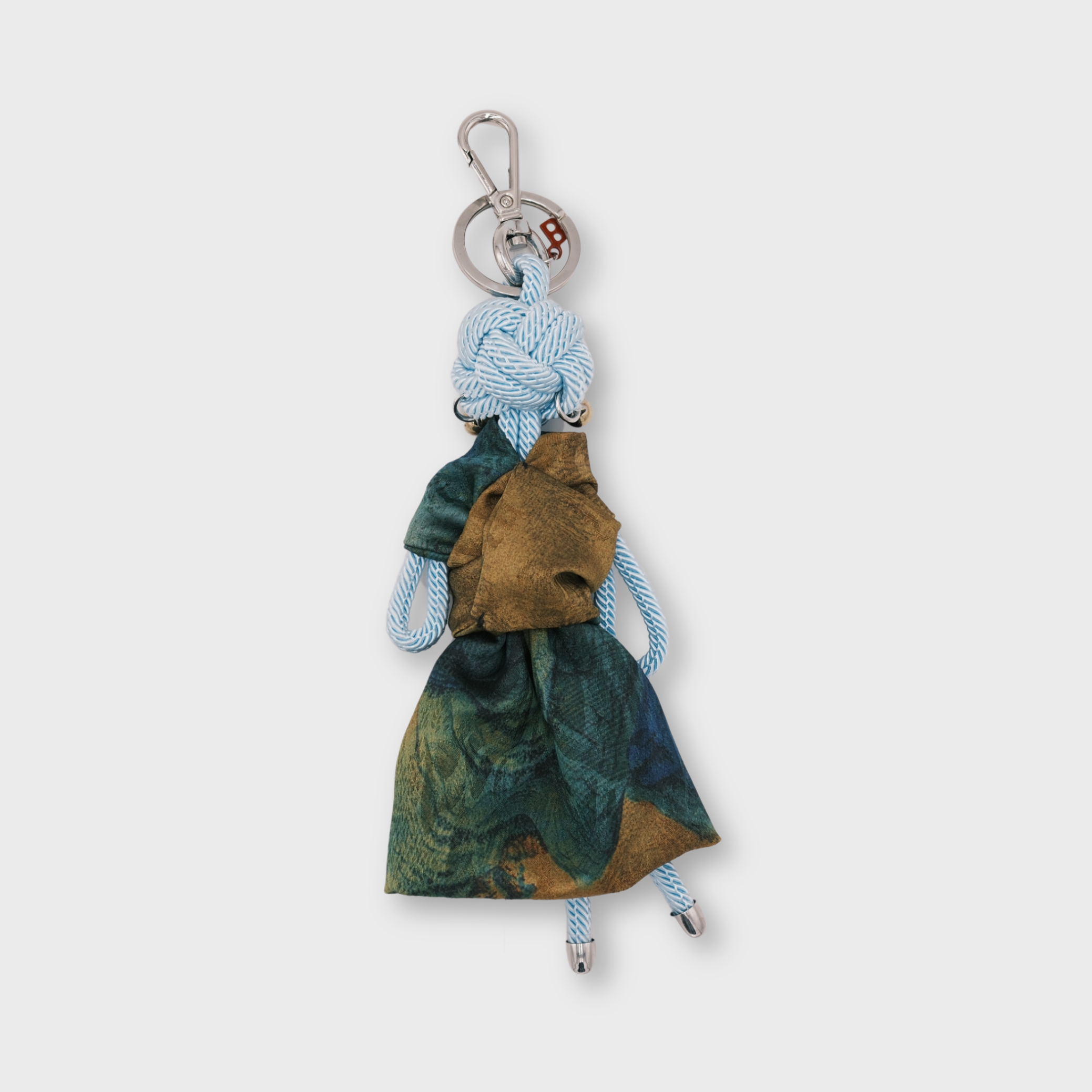 A handmade macramé keychain featuring a small light blue rope doll with a green and brown patterned dress, gold coloured earrings, and a silver metal key ring at the top.