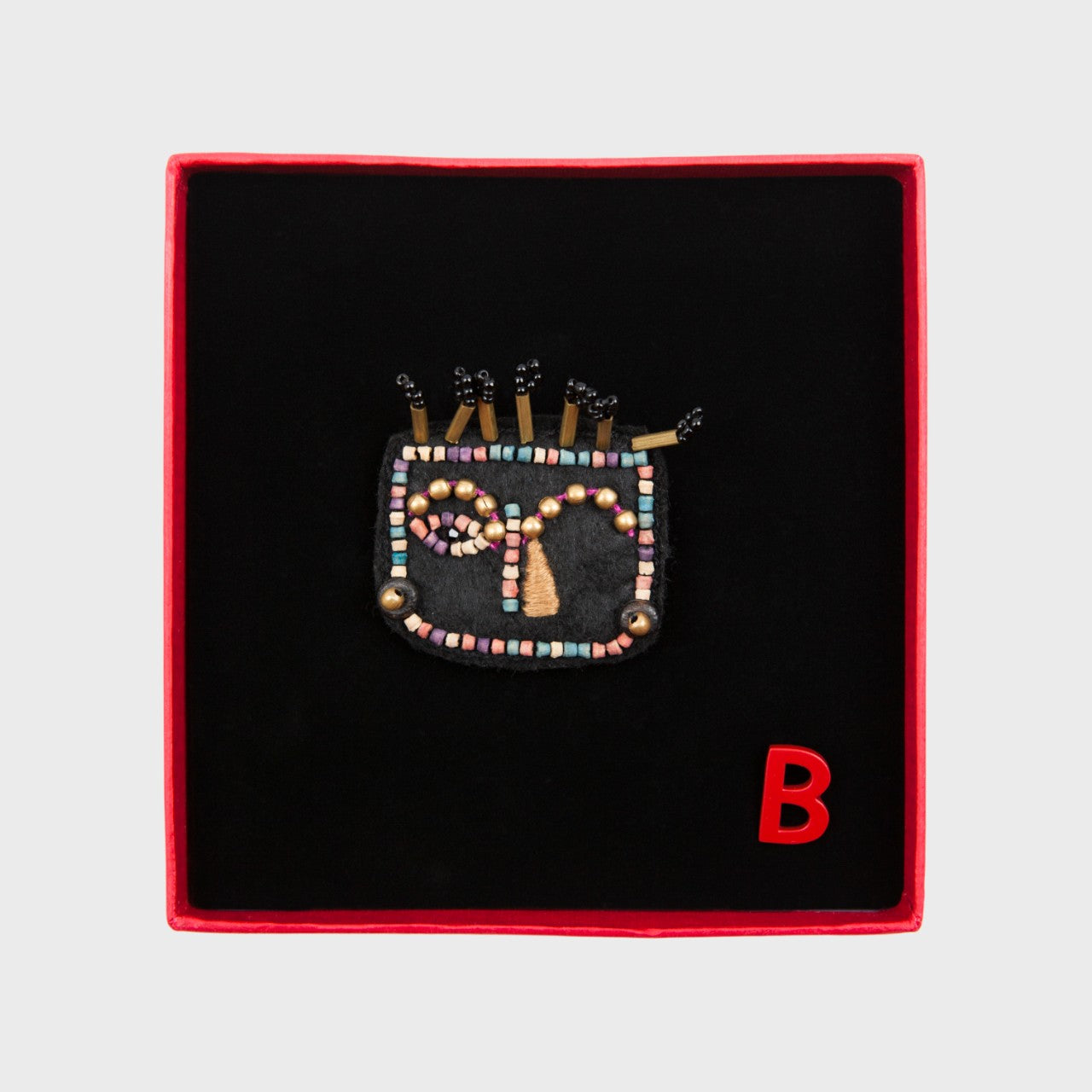 An embroidered brooch representing a face with beige details in a box. 