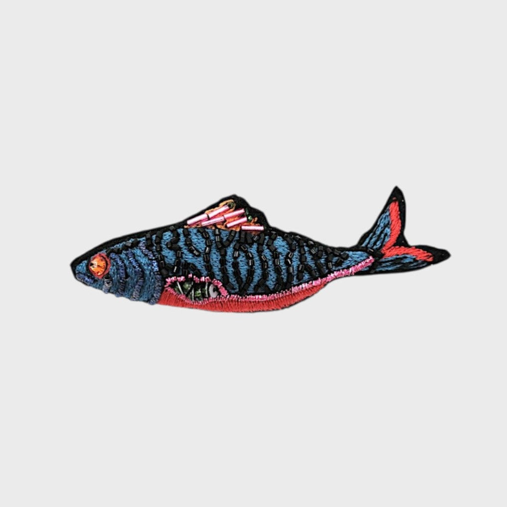 An embroidered blue fish brooch with pink, red and black details. 