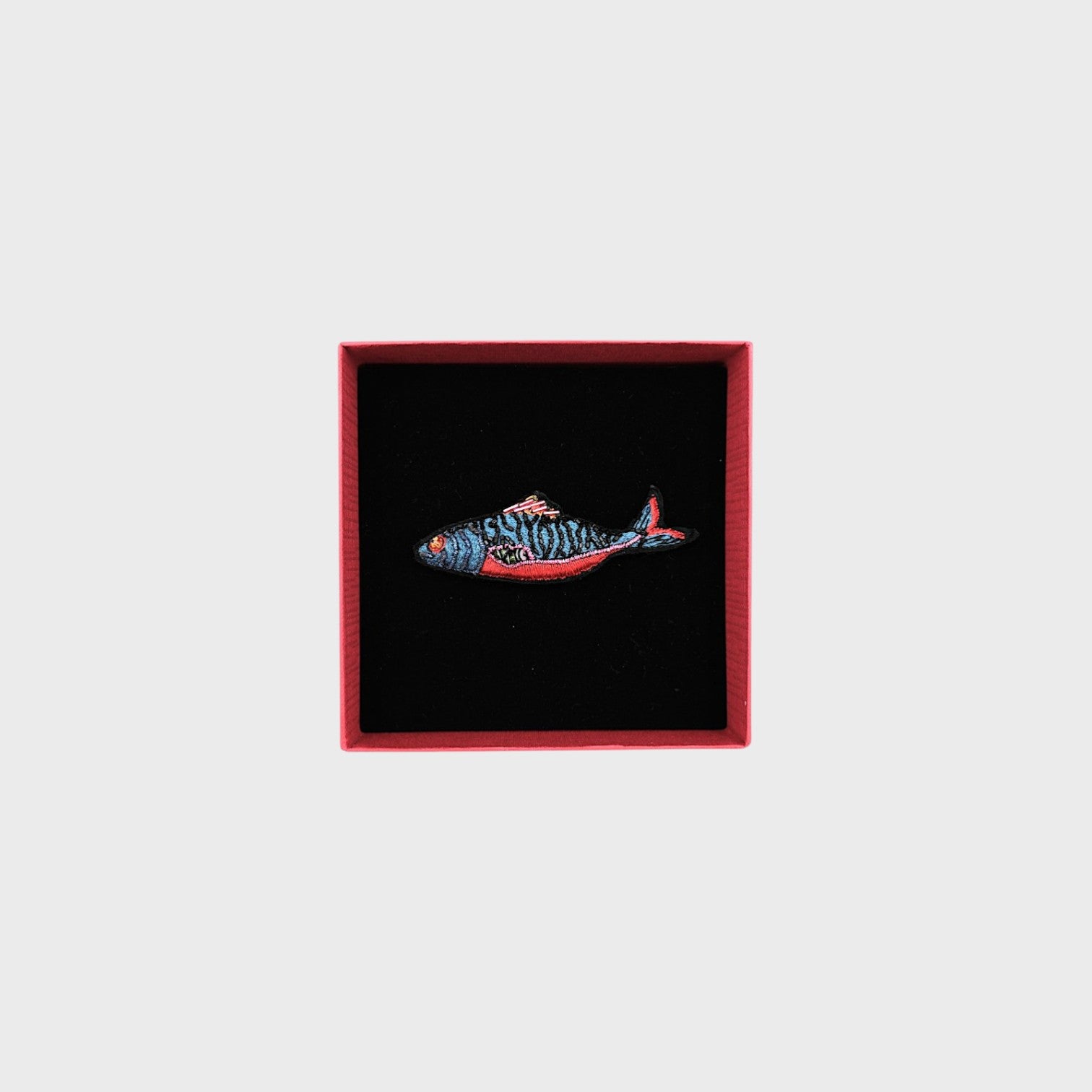 An embroidered blue fish brooch with red, pink and black details in a box. 