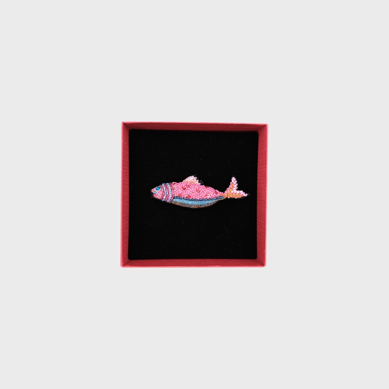 An embroidered pink fish brooch with blue details in a box. 