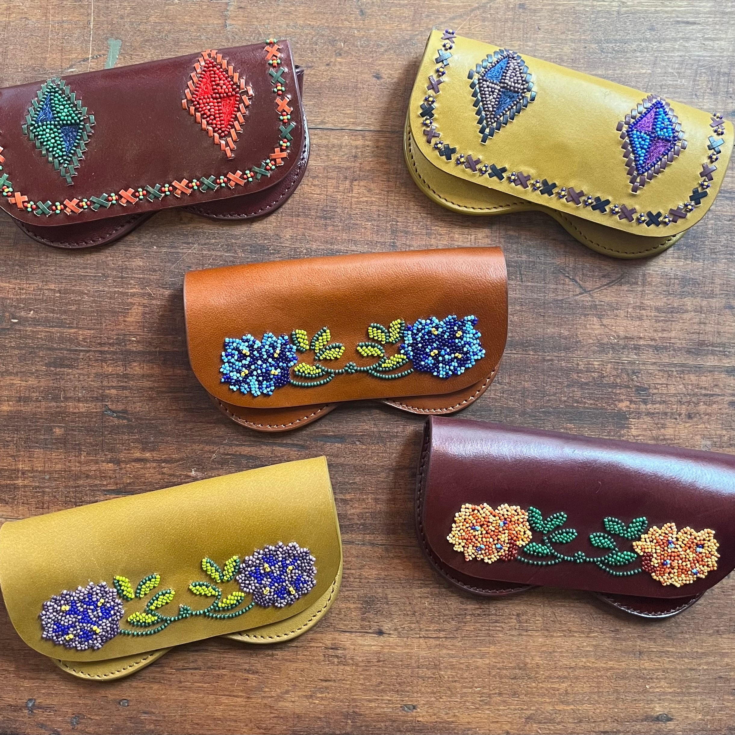 Five leather glasses cases arranged on a wooden surface, including the brown leather flowers one, put in the middle. 