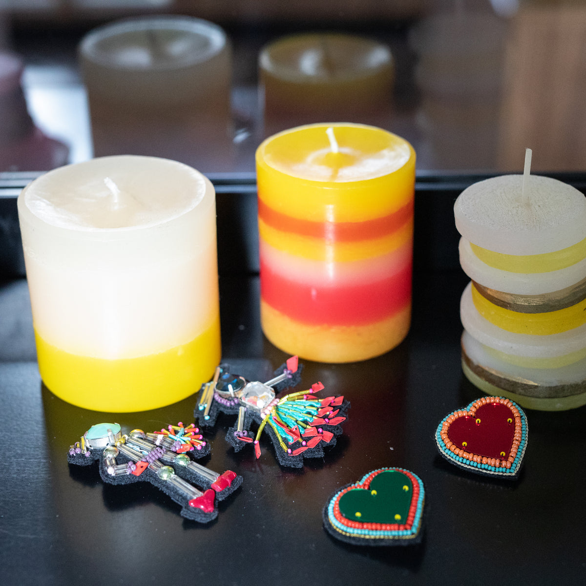 This image features a collection of decorative items arranged on a dark surface, likely a part of a gift set. The items include three uniquely designed candles in different shades of yellow and a striking heart-shaped brooches, all arranged to showcase their aesthetic appeal.