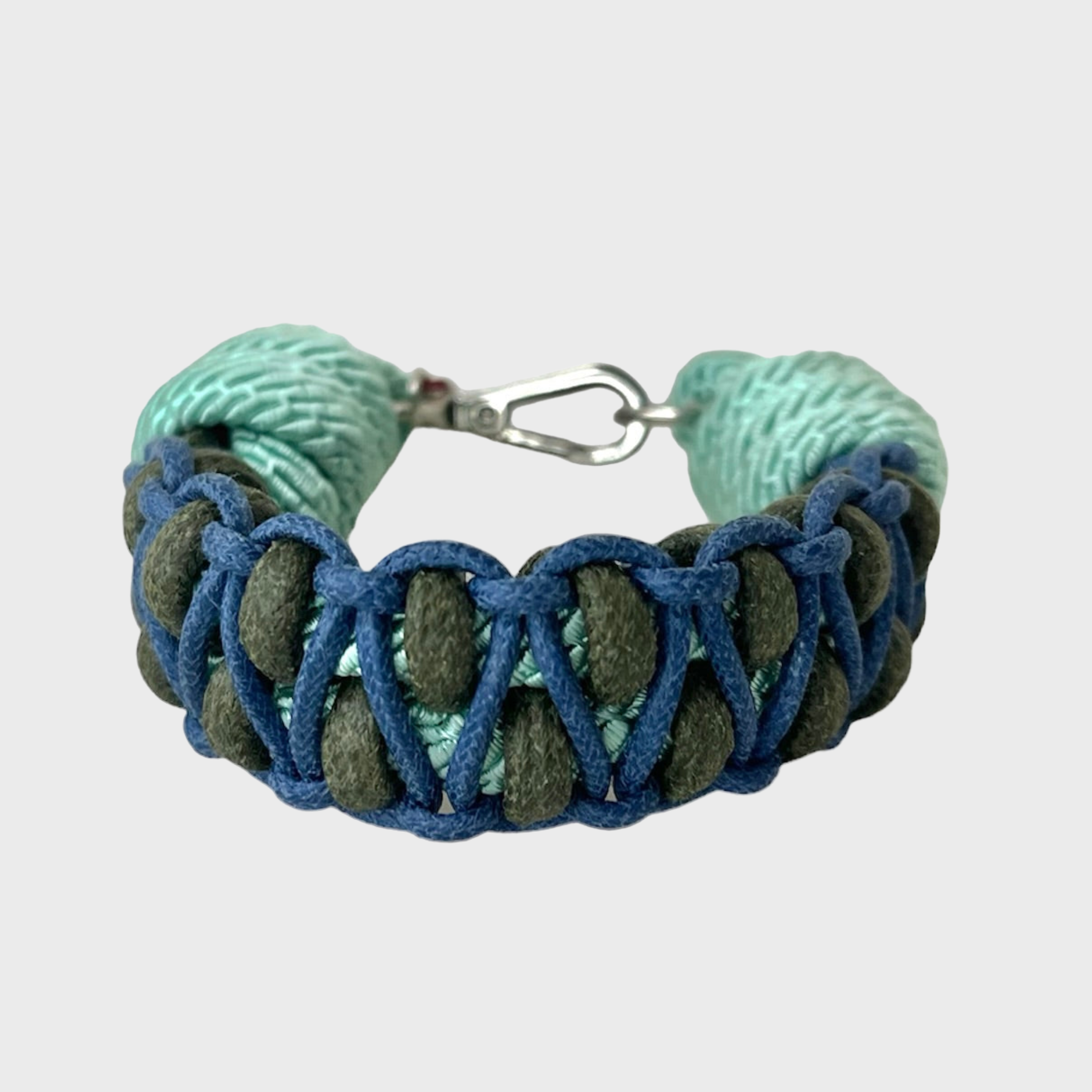 A braided macramé bracelet with shades of dark green, light and dark blue. It ends and a silver clasp, shown on a light gray background.
