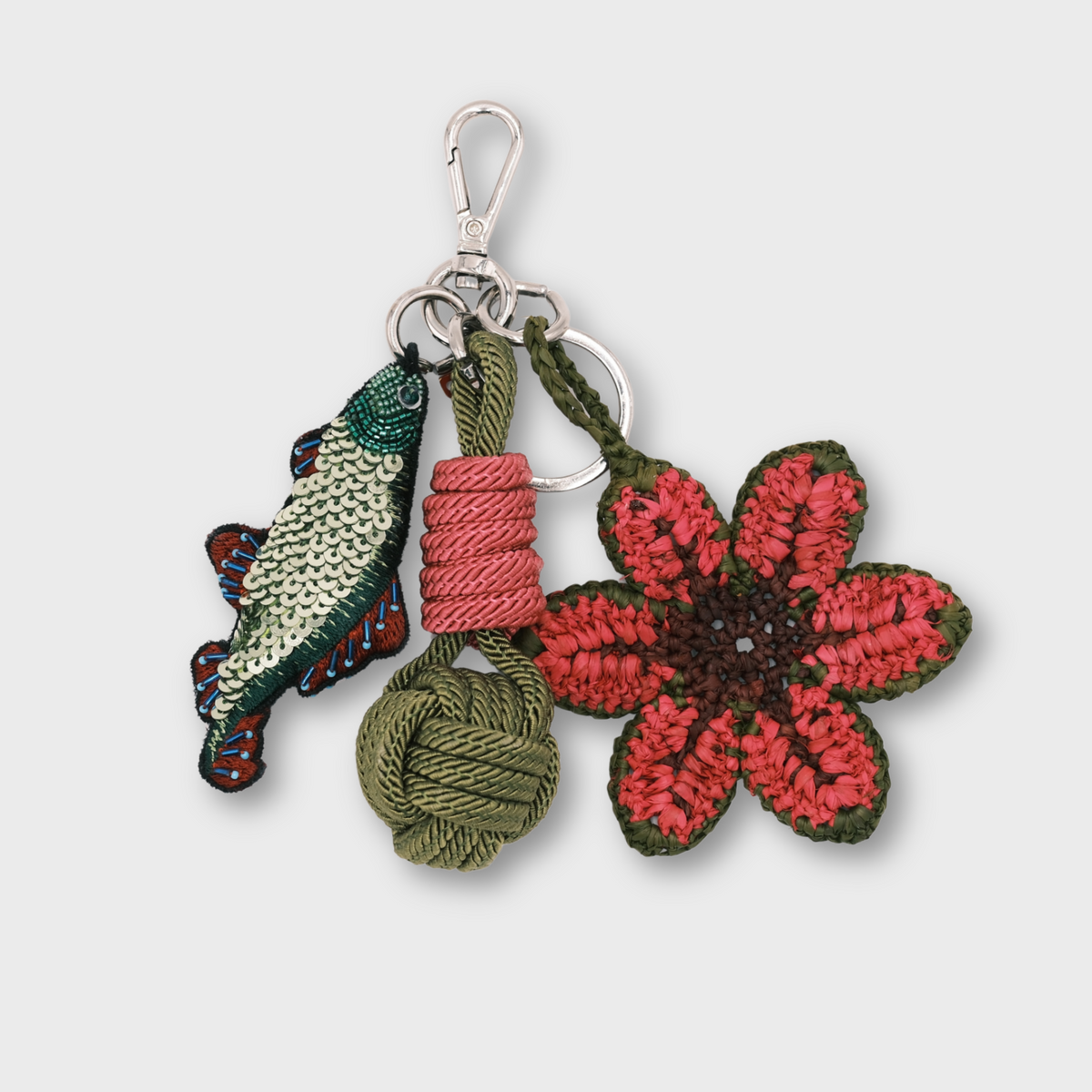 A small embroidered keychain charm set with a silver clasp, featuring a pink and green flower, a green fish, a pink wrapped cord, and an olive green knotted cord on a light gray background.