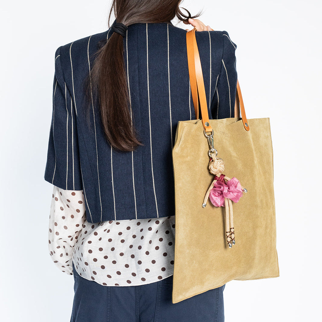 A woman shown from behind wearing a navy blue pinstripe blazer, a white polka dot shirt, and navy trousers, carrying a large green suede tote bag with leather straps and a pink ballerina keychain.