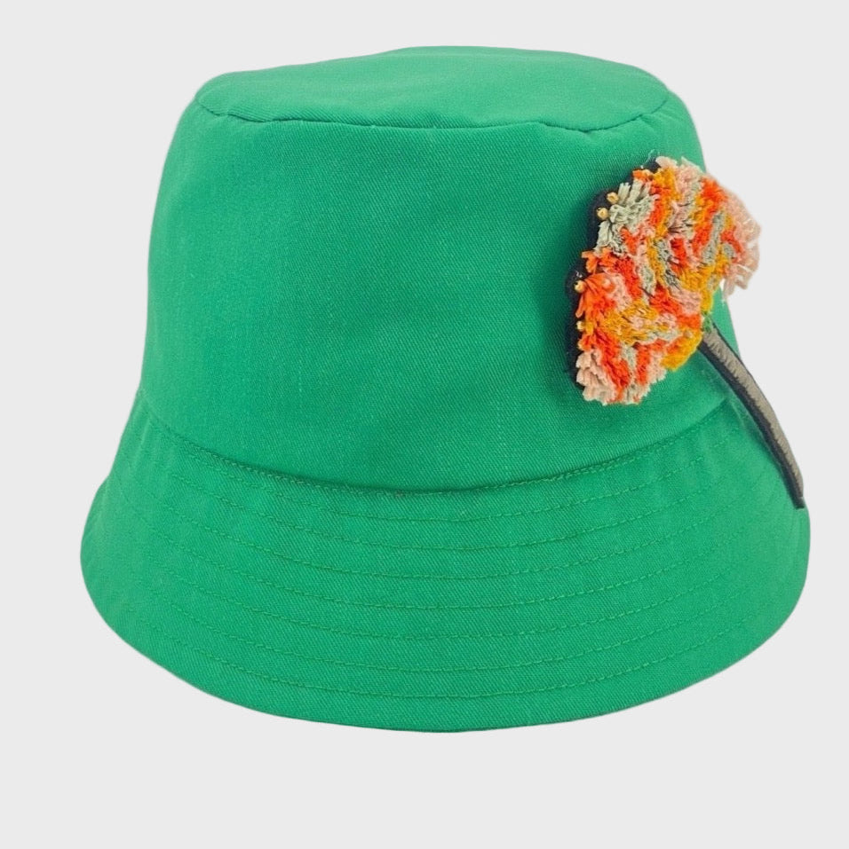 A green cotton bucket hat with an orange carnation flower brooch pinned on it. 