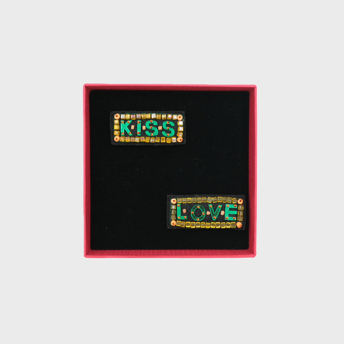 Two decorative brooches with 'KISS' and 'LOVE' text in a box on a light gray background