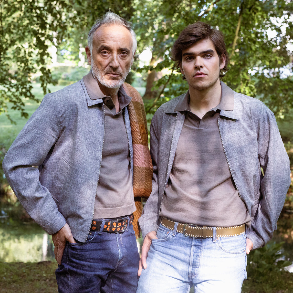 Two men standing outdoors, one wearing the green leather belt with white stitching with trees in the background