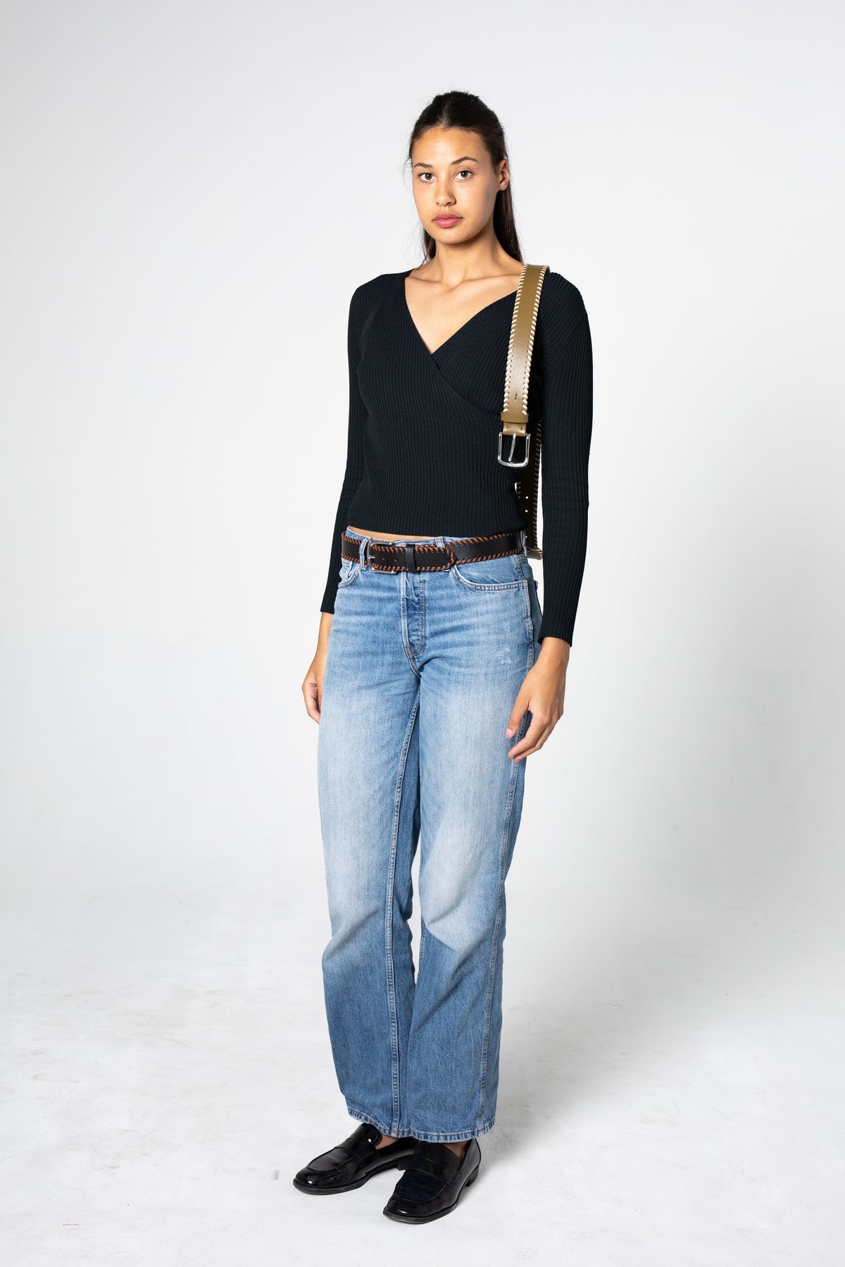Woman wearing a black top, blue jeans, and black shoes with a green leather belt on her shoulder on a white background