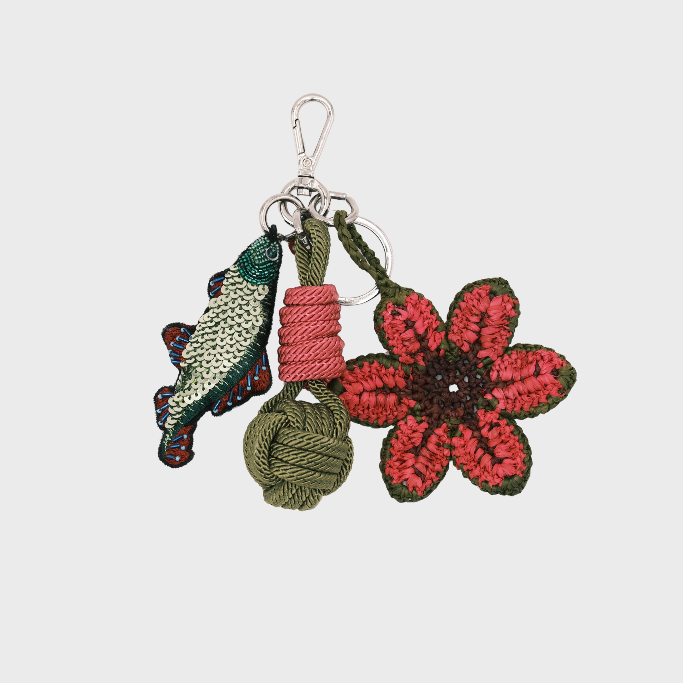 A handmade macramé and raffia keychain featuring a prink crocheted flower, an olive green knotted cord charm, and a green-and-red fish charm, attached to a silver metal clasp.