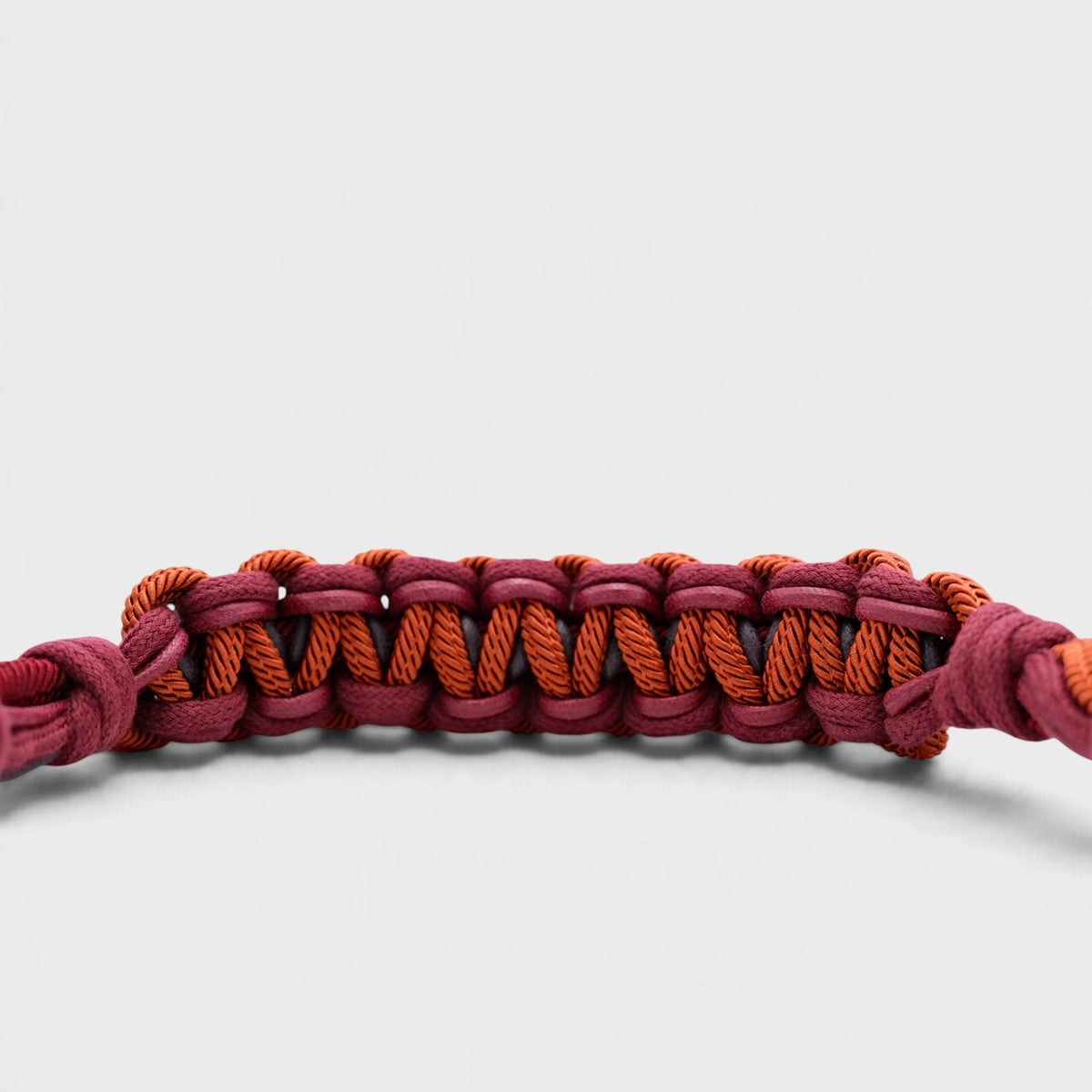 A close-up of a woven macramé bag strap in alternating dark red and orange-brown cords, shown against a light gray background.