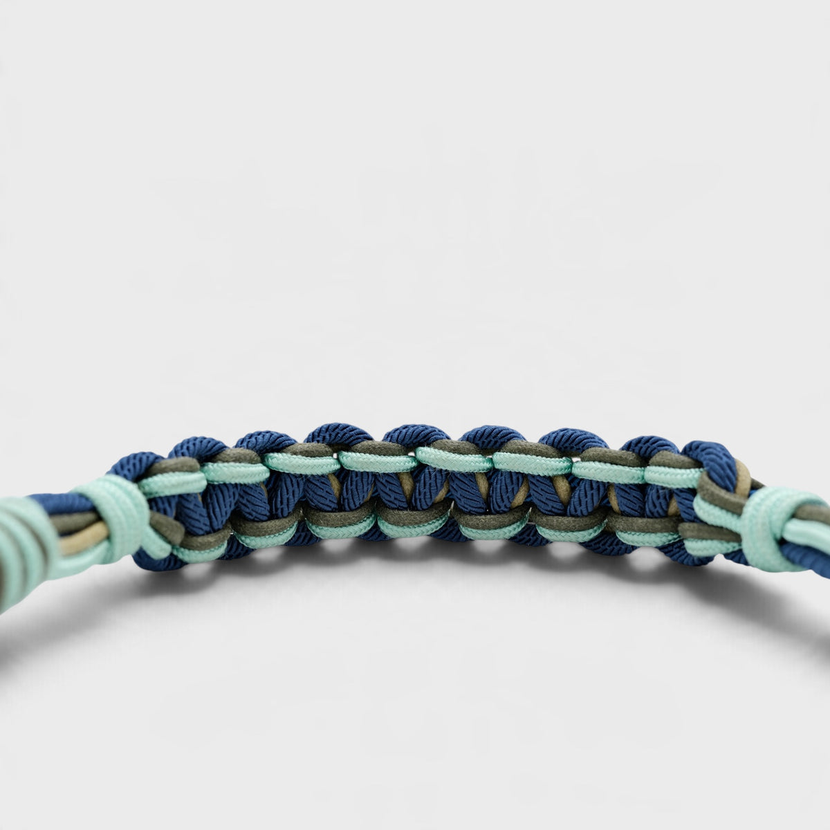 A close-up of a braided macramé bag strap in navy blue, mint green, and olive drab, shown against a clean light gray background.