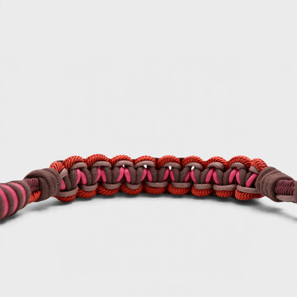 A close-up of a woven macramé bag strap with alternating red and brown braided sections and small pink accents, photographed on a light gray background.