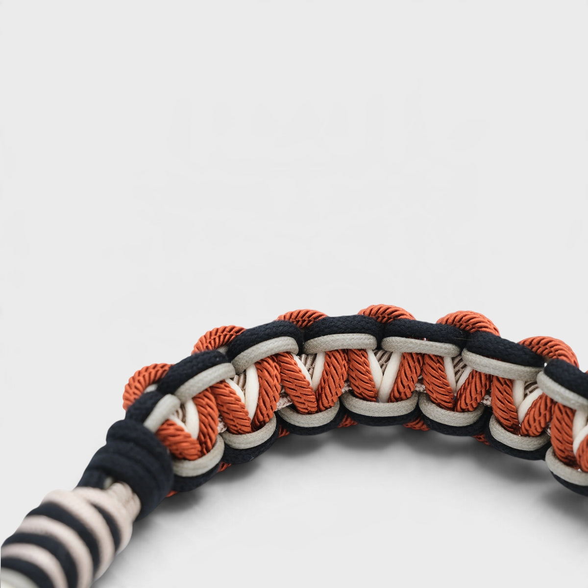 A close-up of a woven macramé bag strap with alternating black and orange loops and white accent lines, photographed on a clean light gray background.