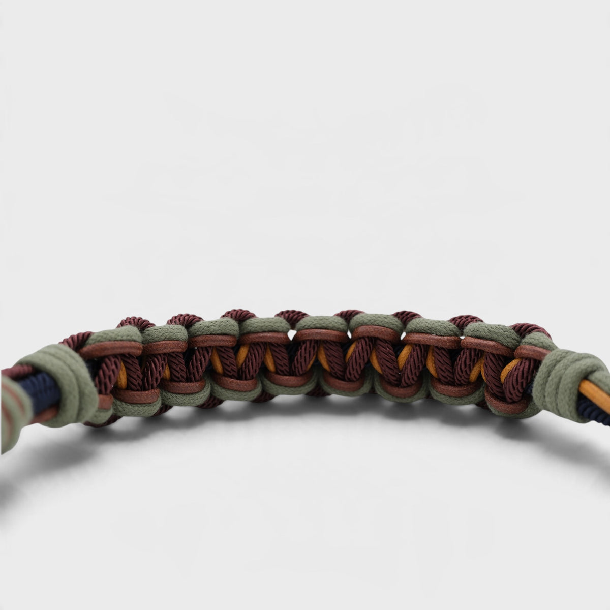 A close-up of a woven macrame shoulder strap, featuring alternating loops of dusty sage green, dark burgundy, and mustard yellow on a light gray background.