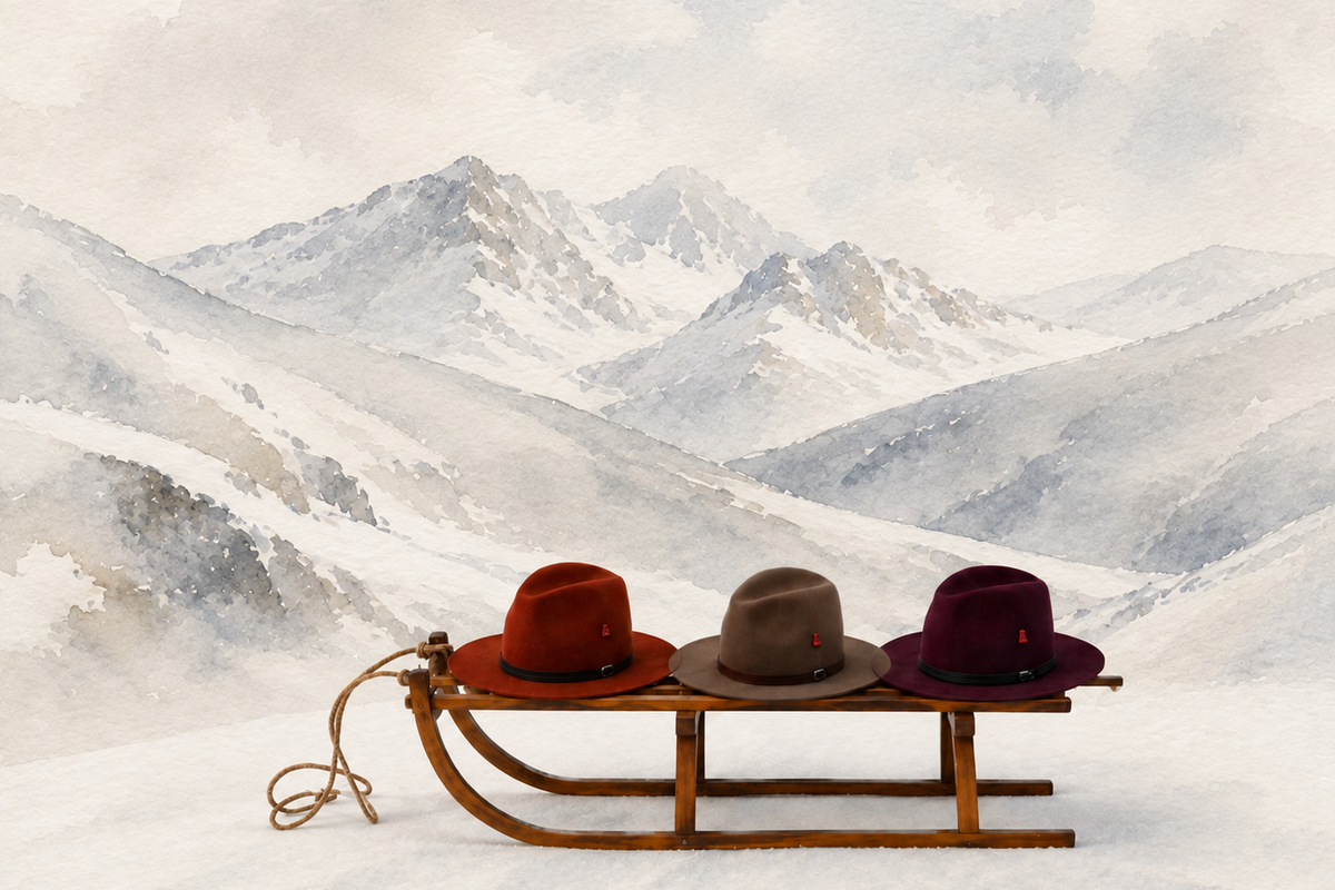 Three wool felt fedora-style hats in rust red, taupe, and plum rest on a wooden sled in a snowy mountain landscape rendered in a soft watercolor style.