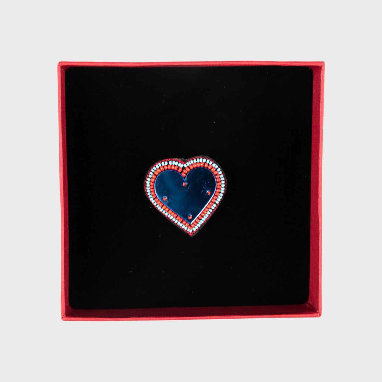 An embroidered brooch representing a heart with a blue glass middle in a box. 