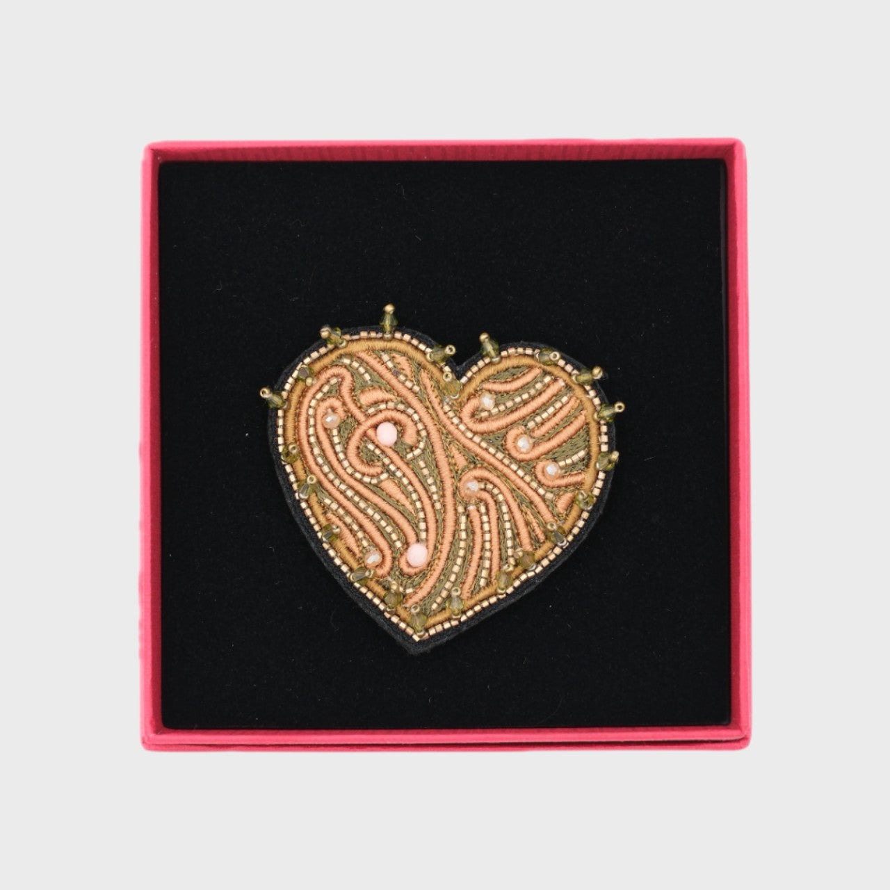 An embroidered brooch representing a heart with green details in a box. 