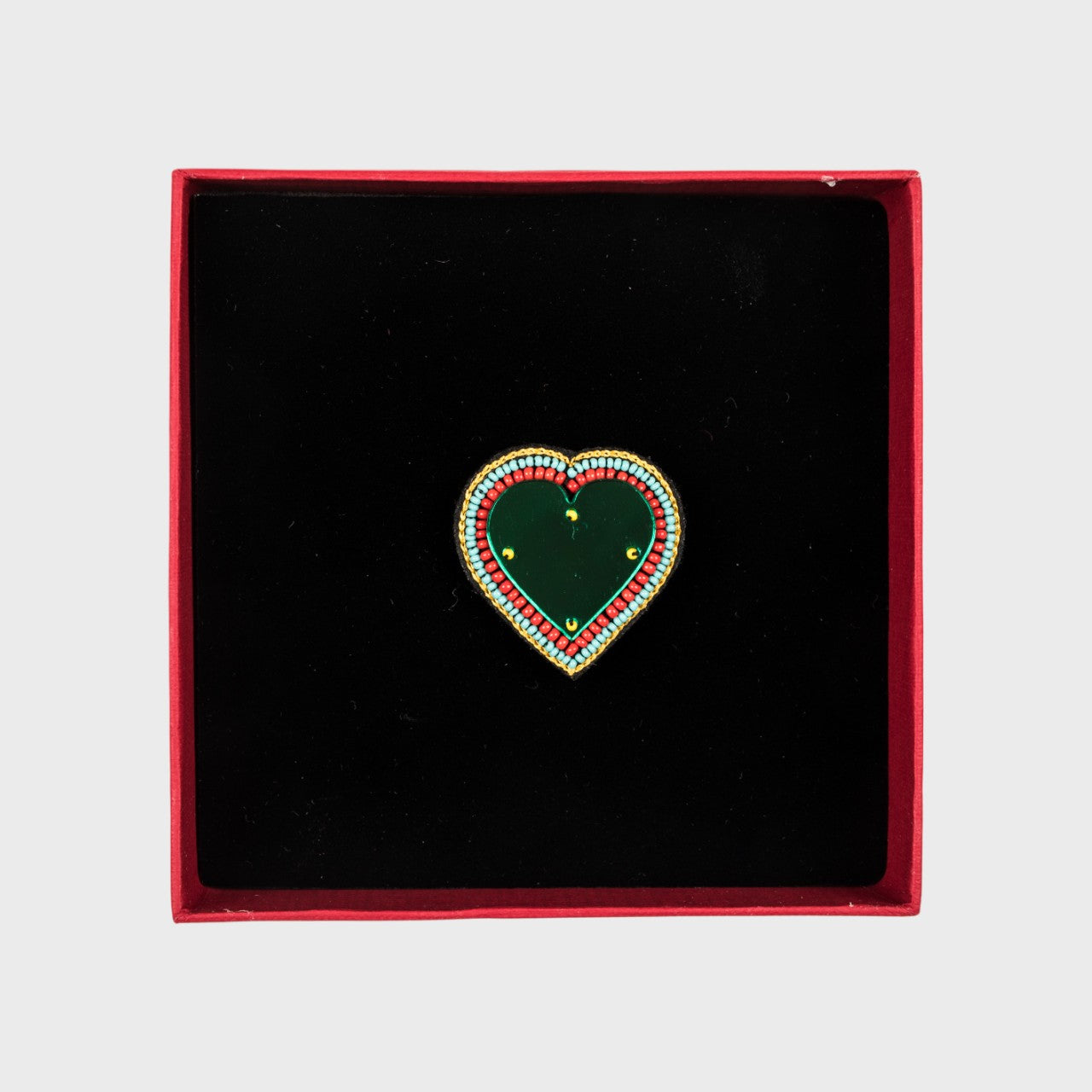 An embroidered brooch representing a heart with a green glass middle in a box. 