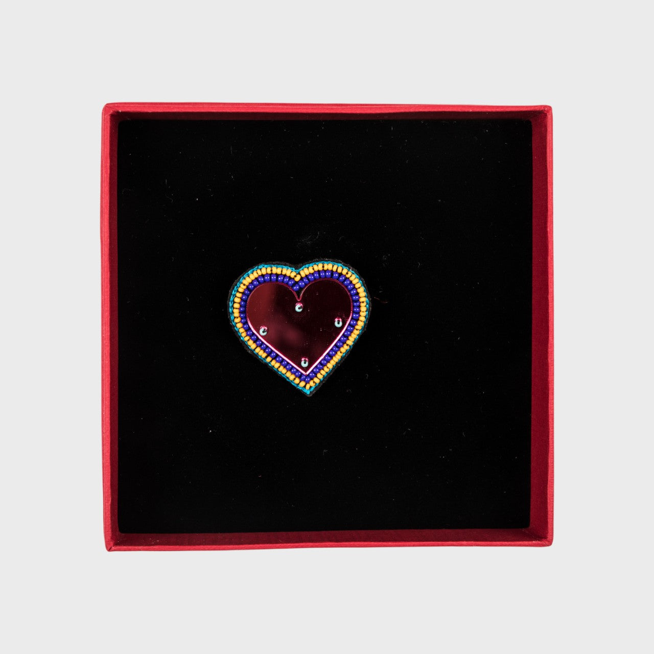 An embroidered brooch representing a heart with a pink glass middle in a box. 