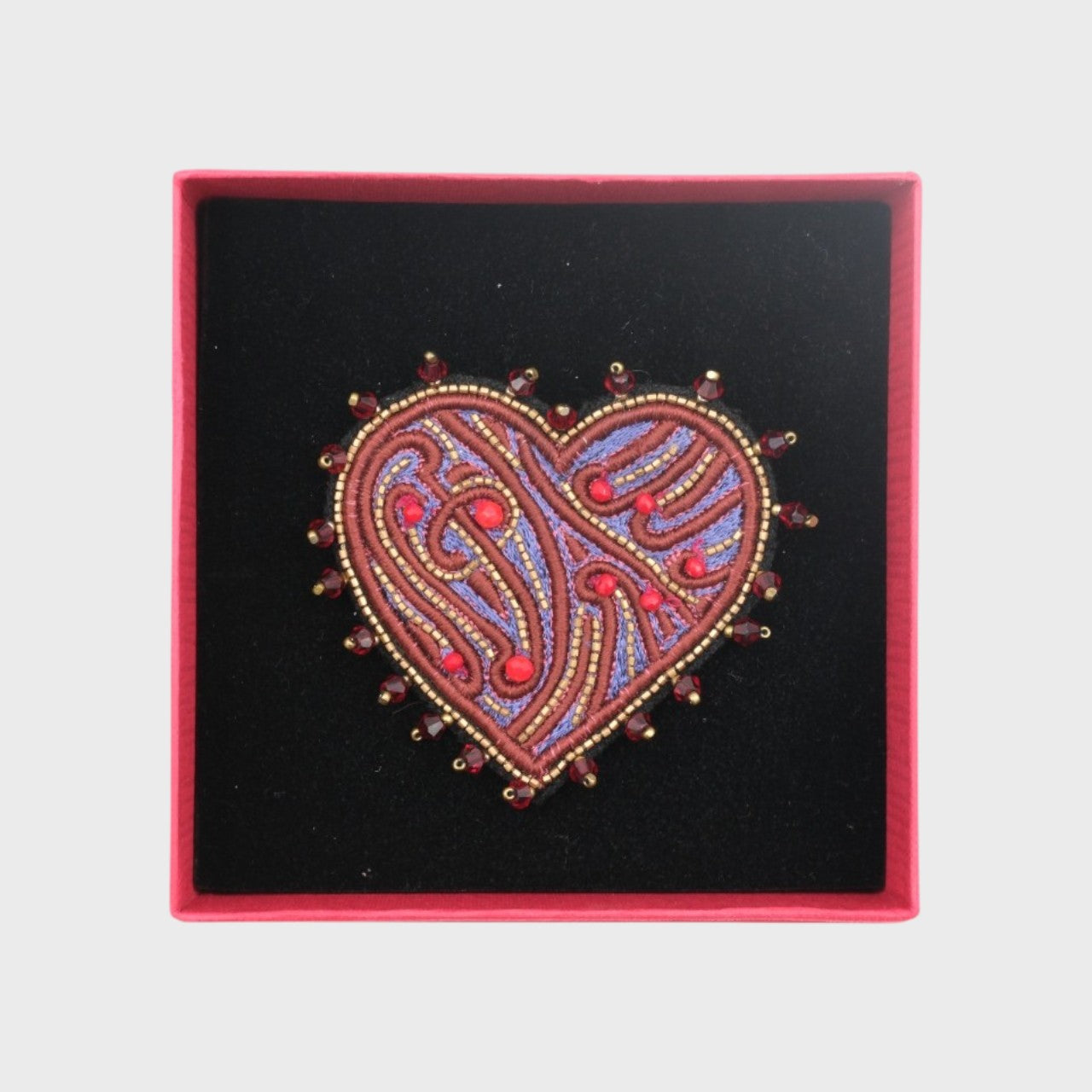 An embroidered brooch representing a heart with purple details in a box. 