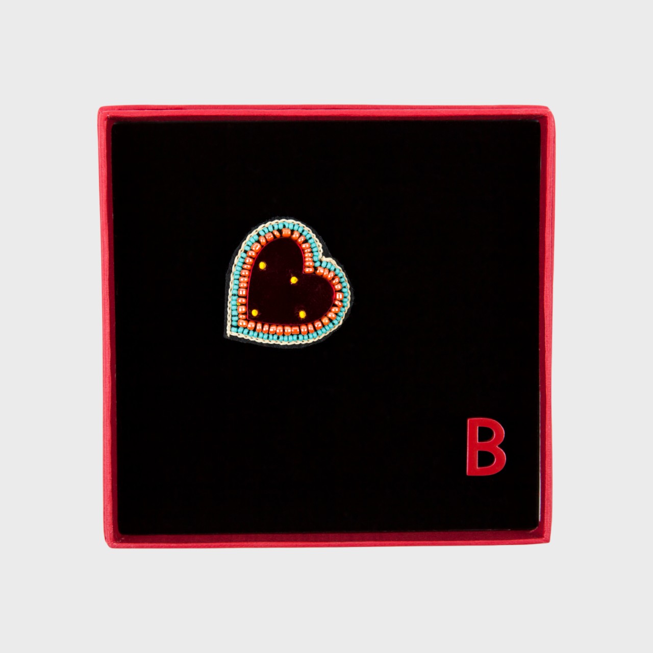 An embroidered brooch representing a heart with a red glass middle in a box. 