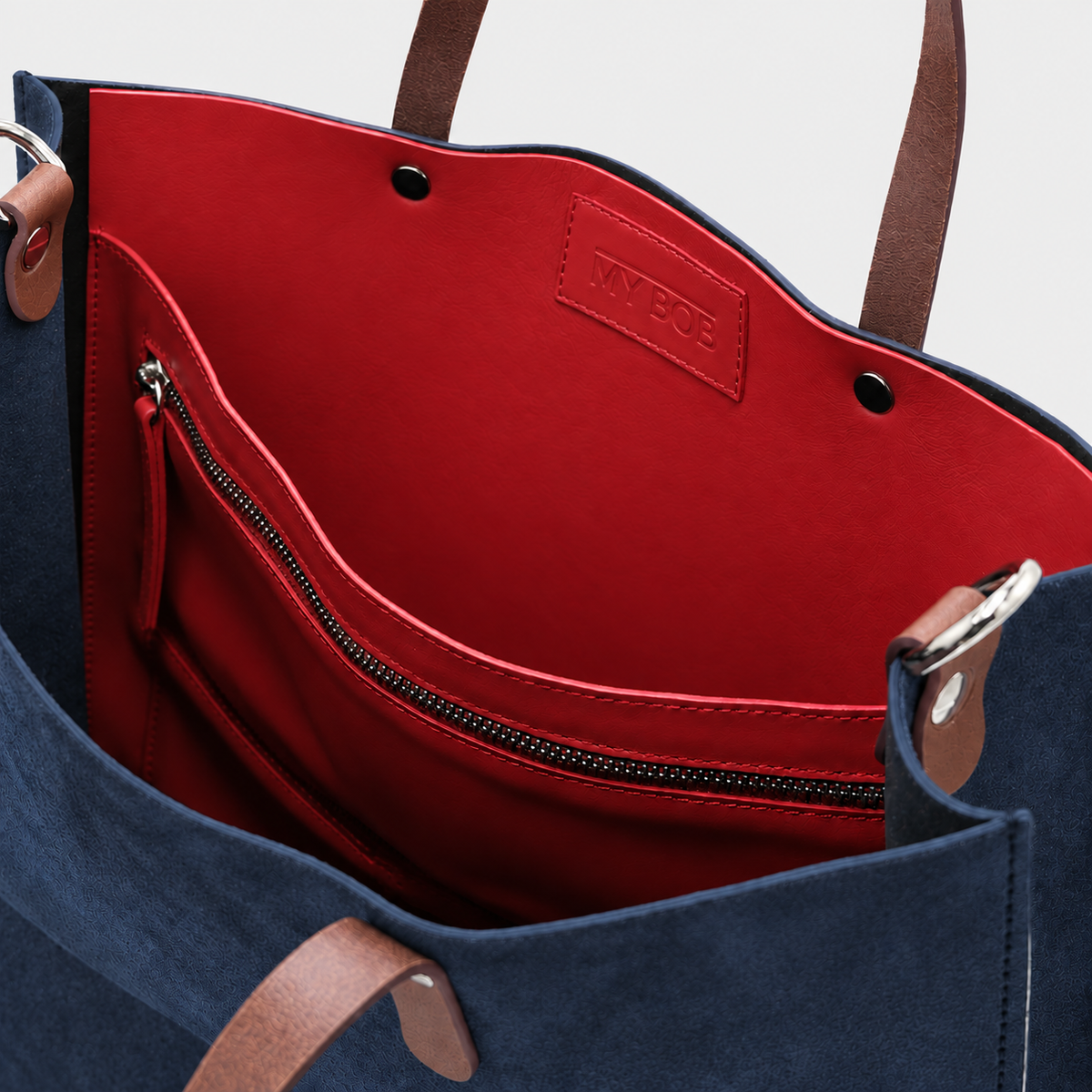 Close-up of the interior of a navy blue tote bag with a bright red lining, brown leather handles, and a zippered pocket. The brand name 'MY BOB' is embossed on a leather patch inside the lining.