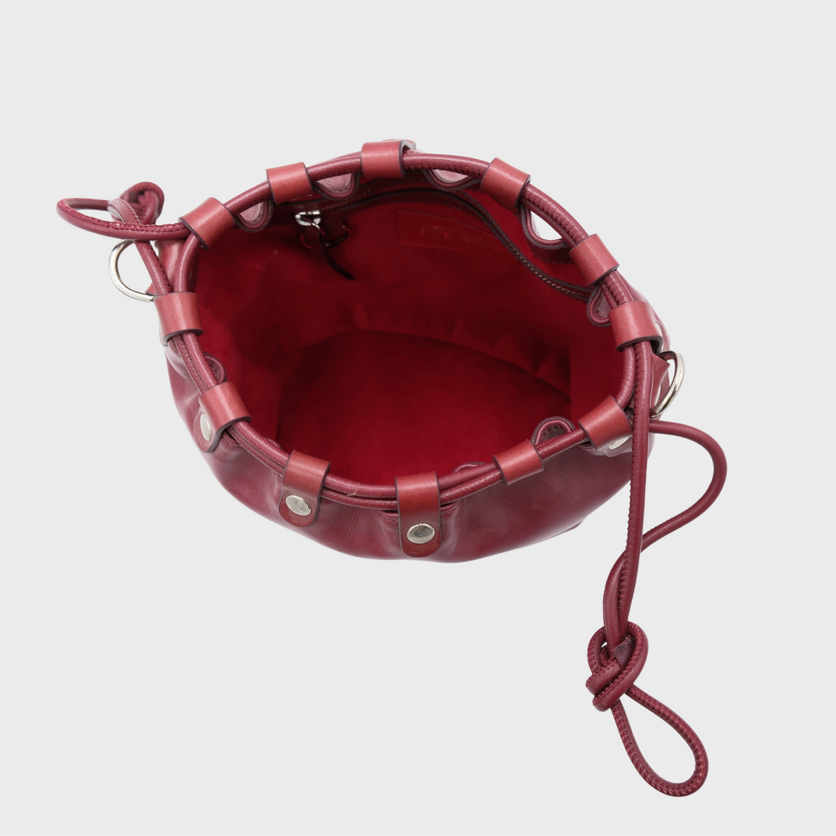 A top-down view of a round, burgundy leather bucket bag with a matching red interior, featuring leather straps around the rim and a knotted leather shoulder strap on a light gray background.